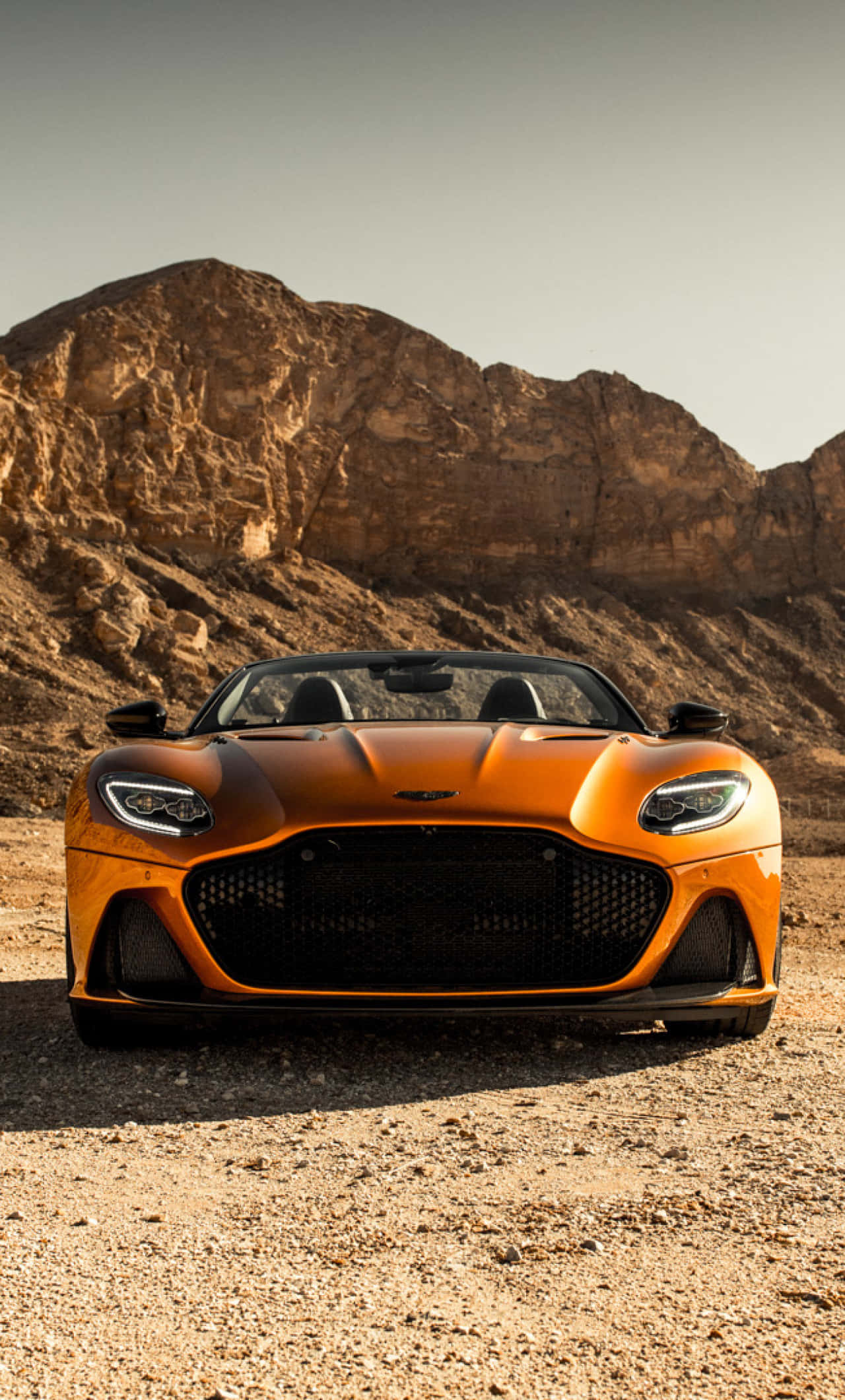 Aston Martin Dbs Superleggera 桌布 壁紙 — 一輛充滿活力的橙色 Aston Martin 跑車停泊在碎石上，背景是崎嶇的沙漠山脈，天空朦朧。
