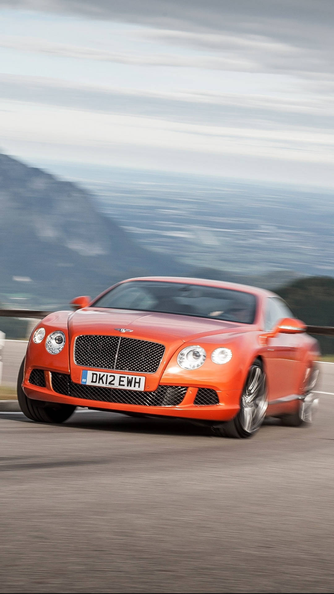 手機 桌布 壁紙 — 一輛充滿活力的橙色 Bentley Continental GT 在模糊的風景和多雲的天空下沿著山路疾馳。