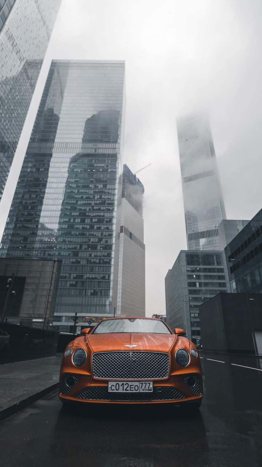 expensive 桌布 壁紙 — 一輛充滿活力的橙色 Bentley 跑車停放在潮濕的城市街道上，下方是高聳入雲、籠罩在薄霧中的玻璃摩天大樓。