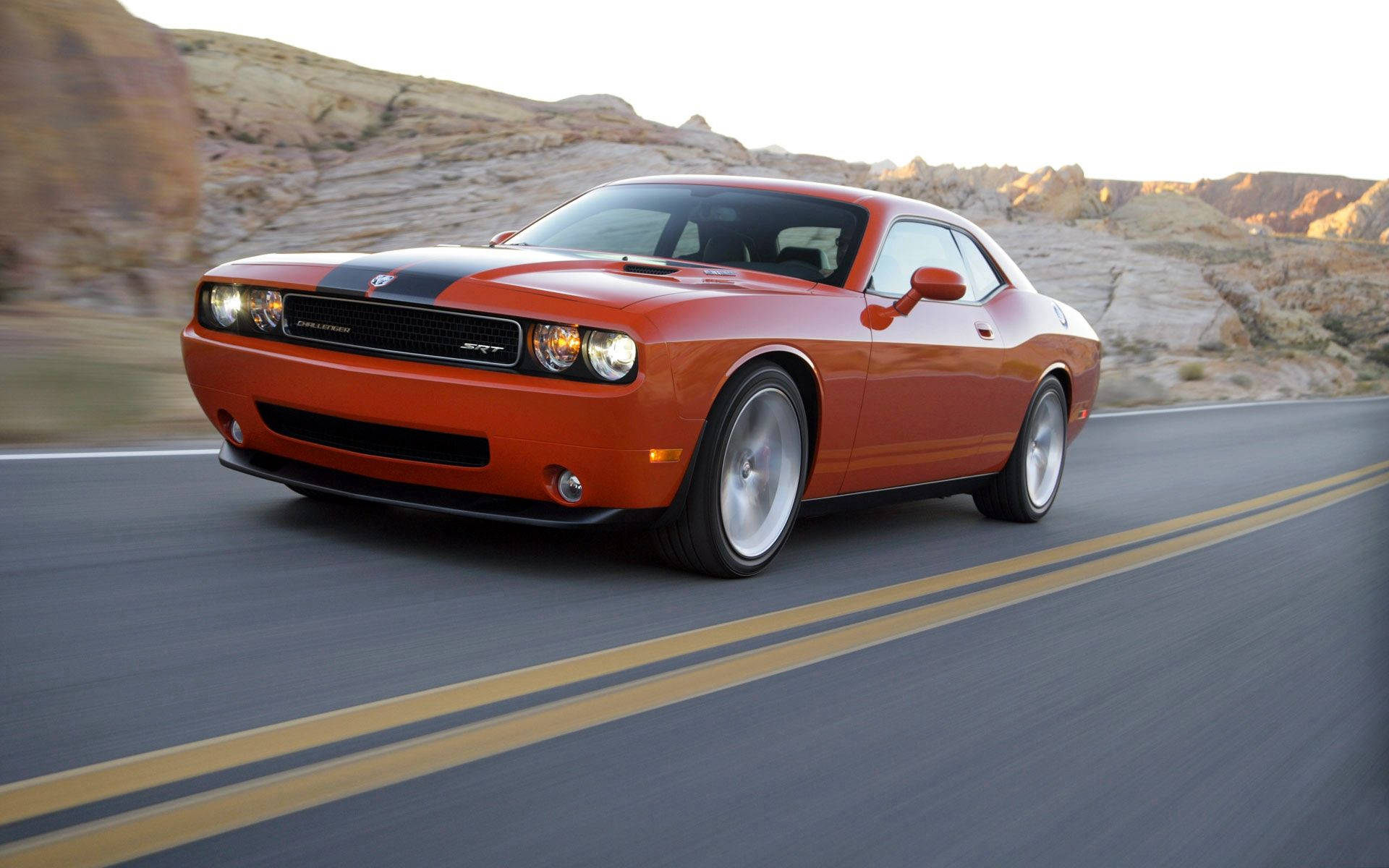 道奇隊 桌布 壁紙 — 一輛充滿活力的橙色 Dodge Challenger 在沙漠公路上疾馳，背景是岩石地形。