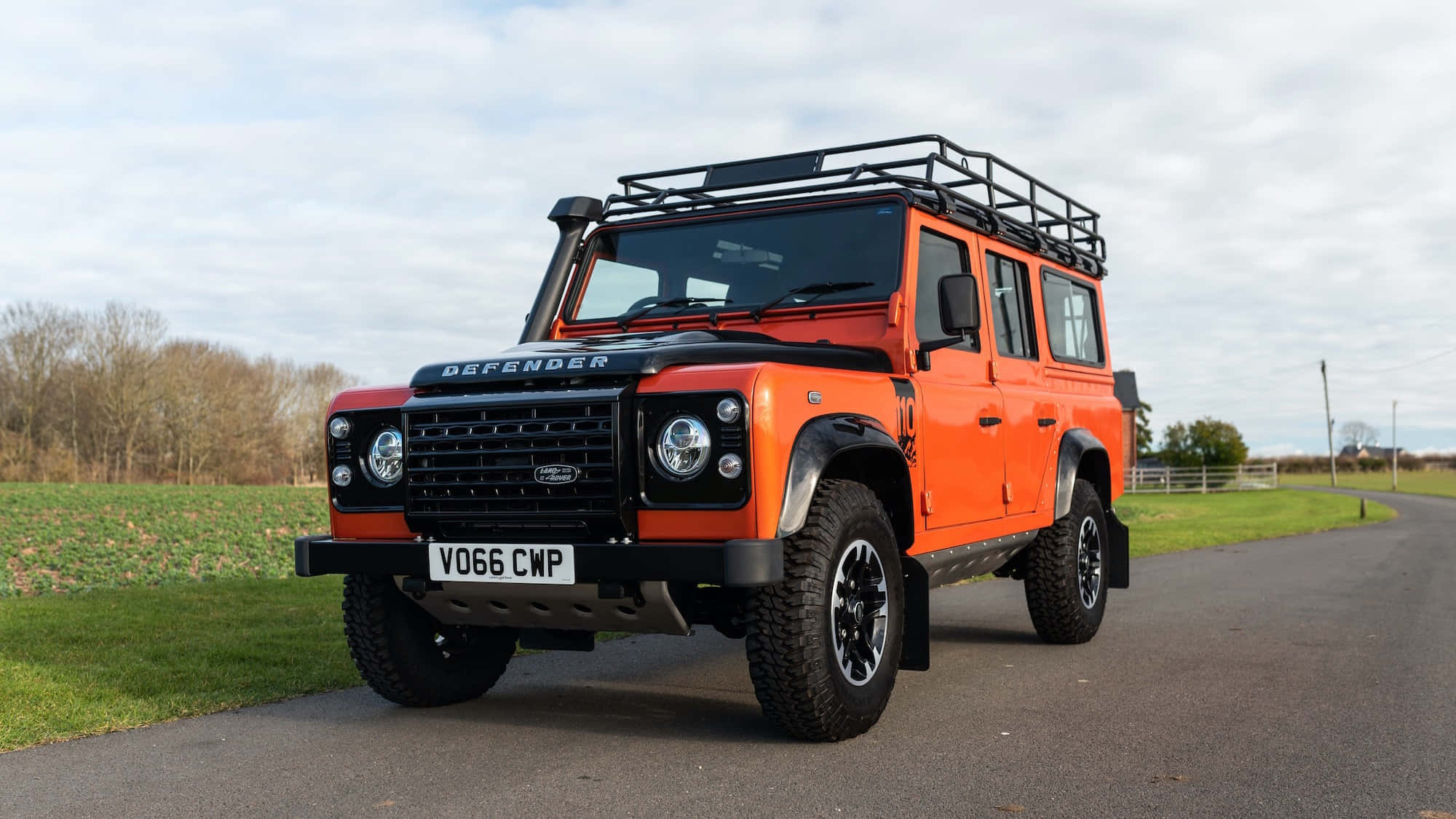 Land Rover Defender 桌布 壁紙 — 一輛充滿活力的橙色 Land Rover Defender，配有黑色車頂行李架，停泊在鄉村道路上，旁邊是綠色的田野，天空多雲。