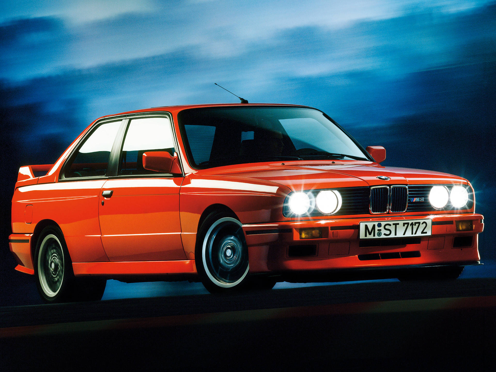 m 桌布 壁紙 — 一輛充滿活力的橙色BMW M3跑車，車頭燈閃爍，在模糊的藍色暮色天空中疾馳。