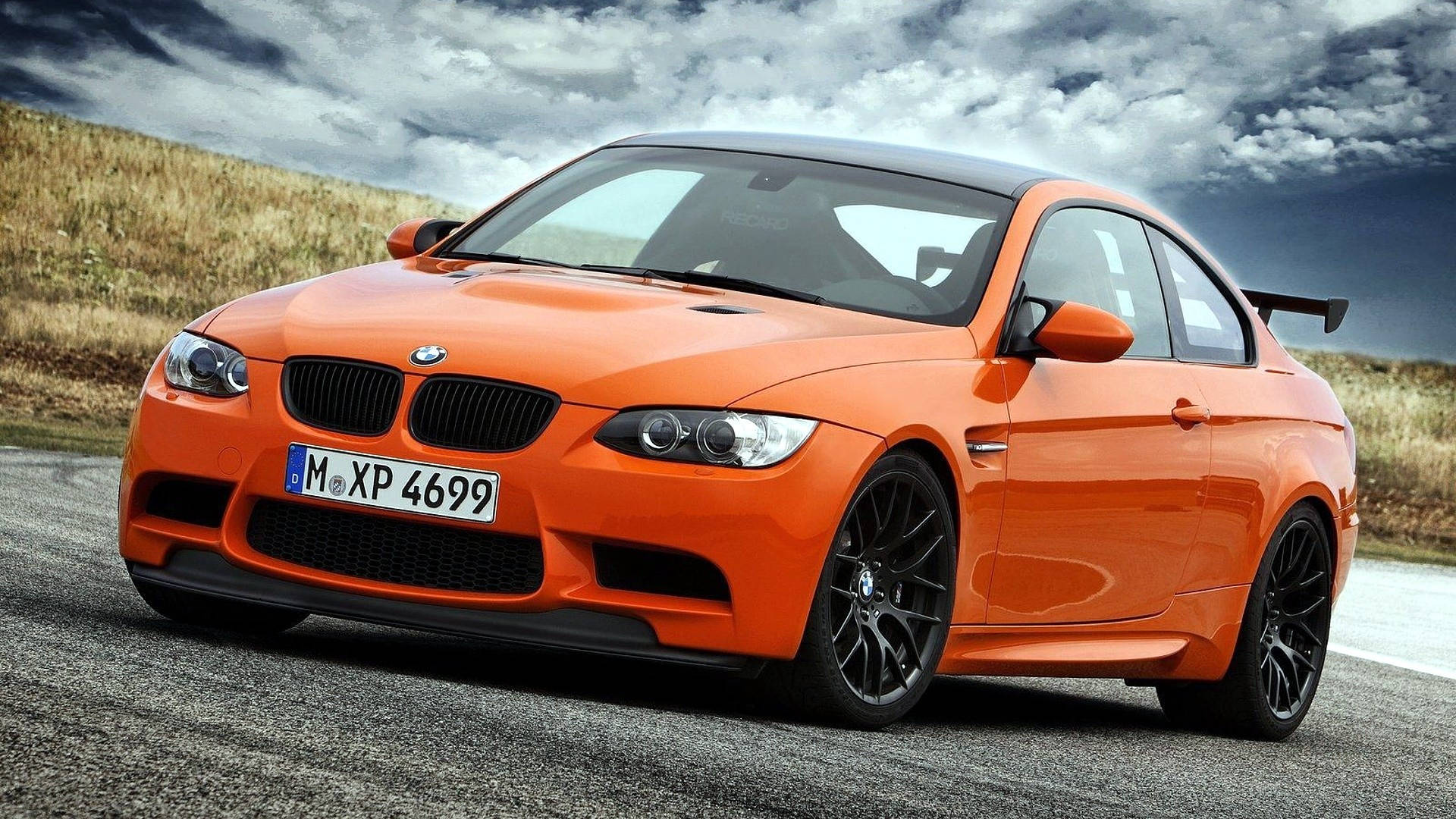 m 桌布 壁紙 — 一輛充滿活力的橙色BMW M3雙門轎跑車，帶有黑色飾件和後擾流板，停在碎石路上，天空烏雲密佈，氣氛戲劇化。