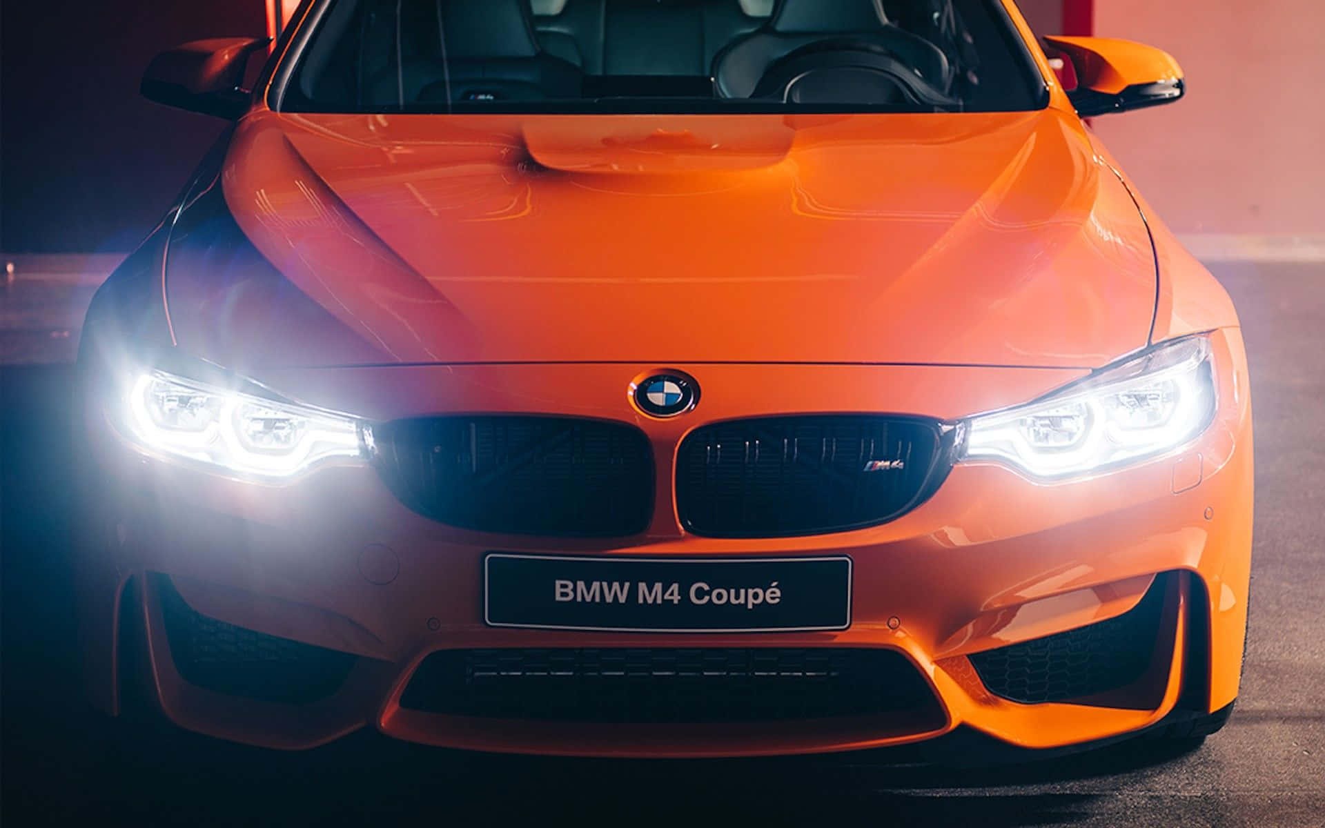 Bmw Cars 桌布 壁紙 — 一輛充滿活力的橙色BMW M4雙門轎跑車在昏暗的環境中，車頭燈閃爍，在戲劇性的燈光下閃閃發光。