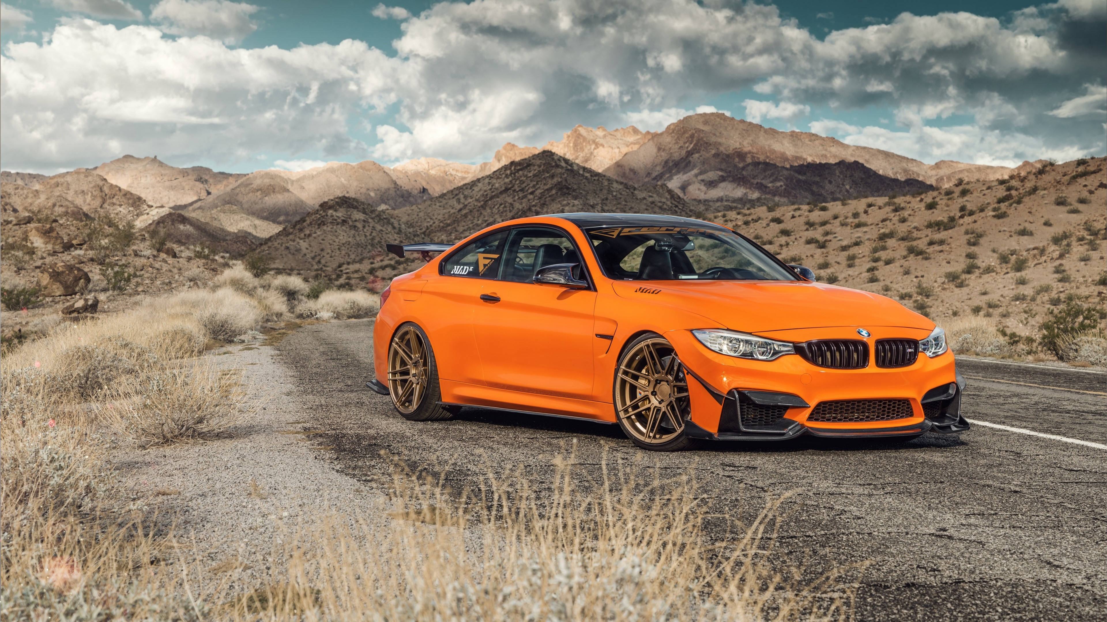 4K BMW 壁紙 桌布 壁紙 — 一輛充滿活力的橙色BMW M4雙門跑車配金色輪圈，停泊在沙漠公路上，背景是崎嶇的山脈和陰天。