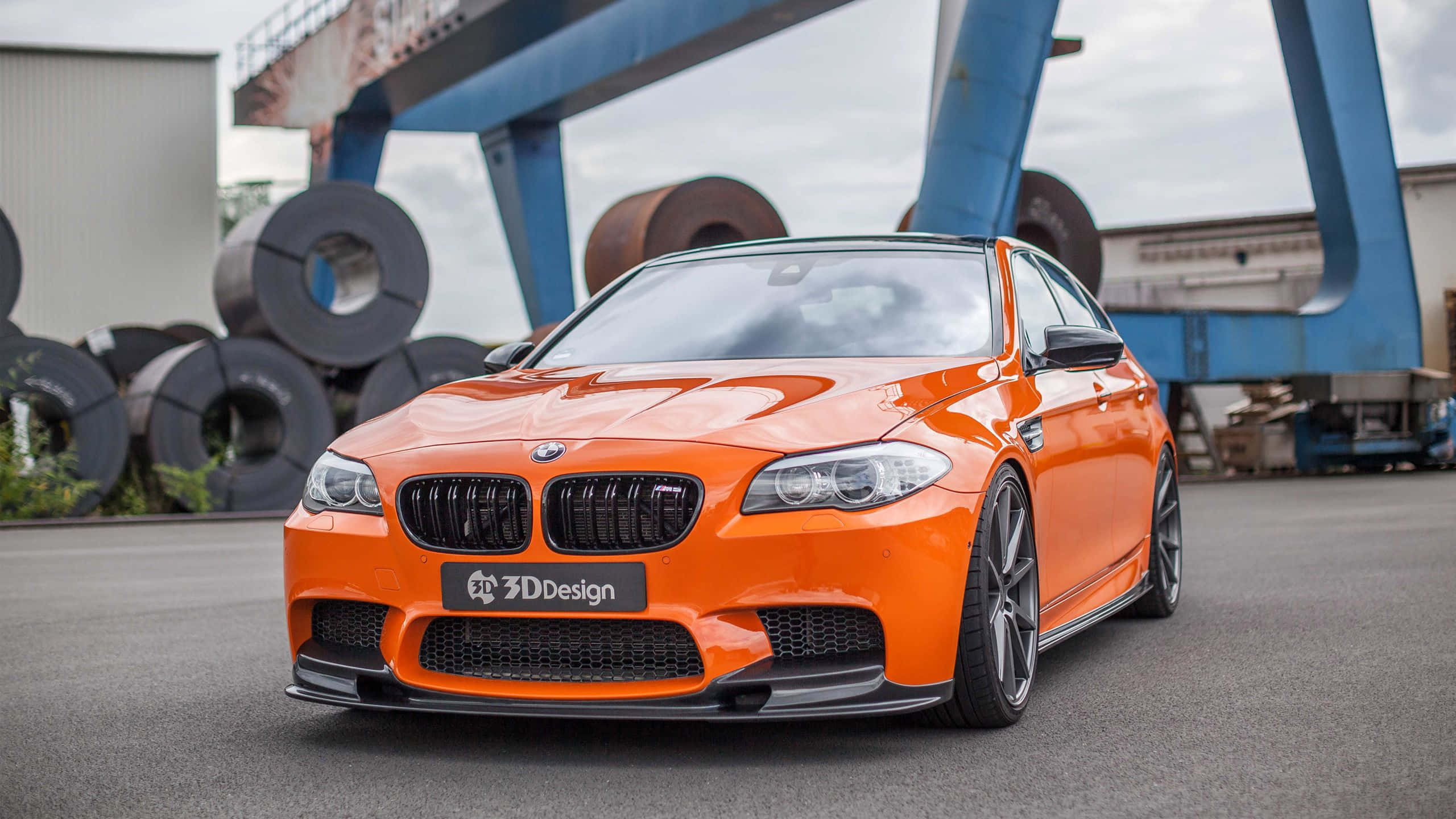 Bmw M5 桌布 壁紙 — 一輛充滿活力的橙色BMW M5跑車，帶有黑色飾邊，停放在工業場地，背景有鋼卷和藍色龍門起重機。
