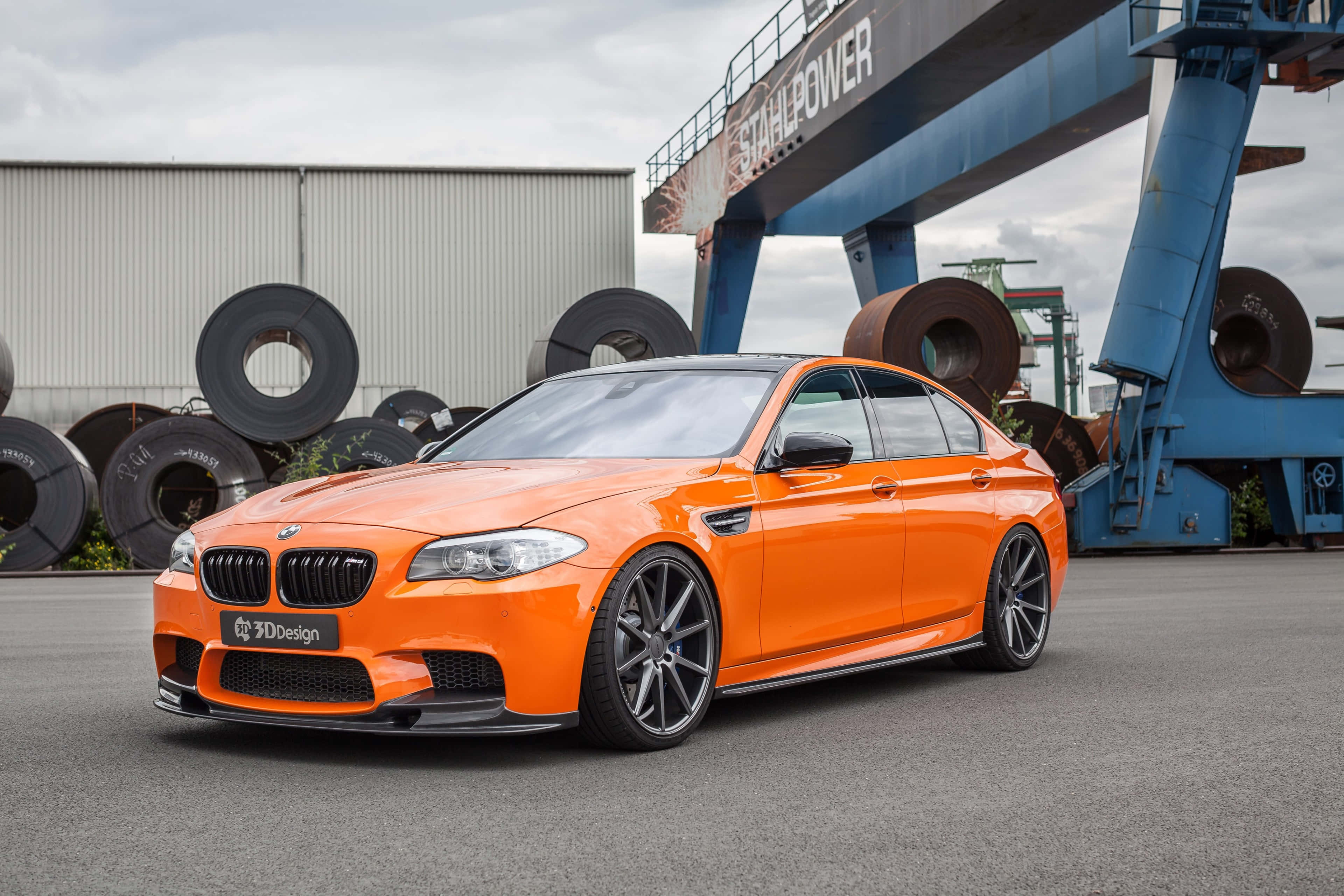 桌布 壁紙 — 一輛充滿活力的橙色BMW M5跑車停放在工業場地，場內有鋼卷和藍色起重機，天空多雲。