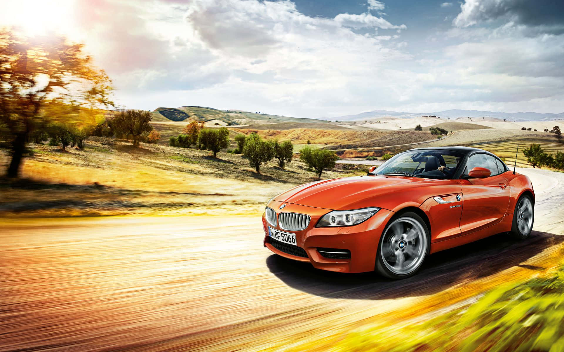 Bmw Z4 桌布 壁紙 — 一輛充滿活力的橙色BMW Z4敞篷跑車在部分多雲的天空下，沿著陽光明媚的鄉村道路穿過金色山丘飛馳。