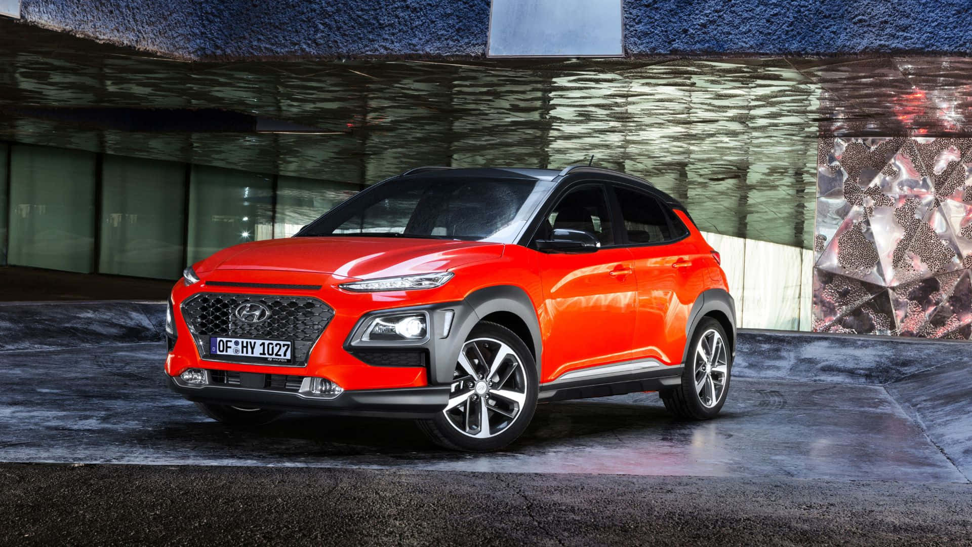 Hyundai Kona 桌布 壁紙 — 一輛充滿活力的橙色Hyundai Kona SUV停在一棟具有紋理牆壁和反光表面的現代建築前。