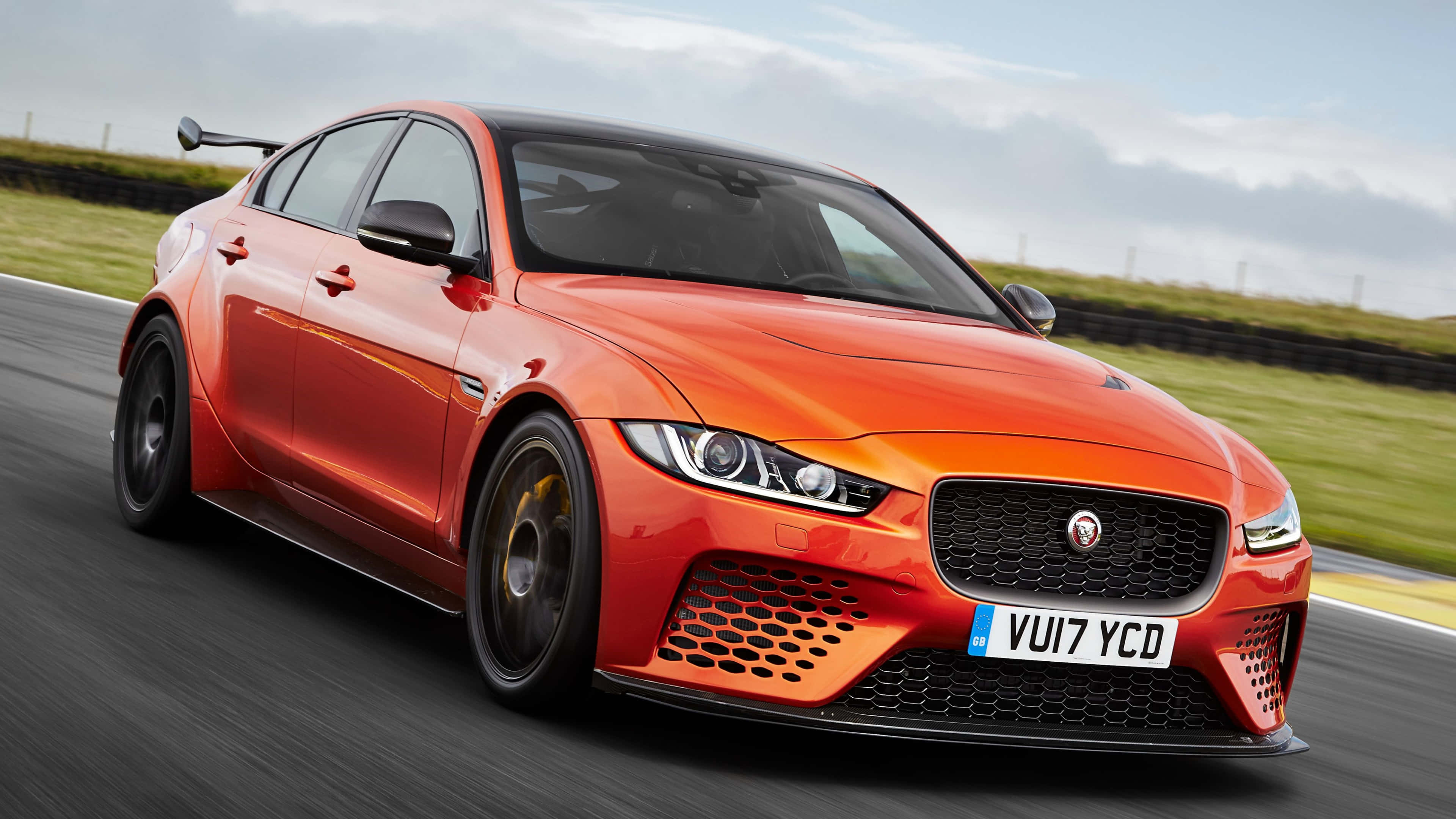 電玩遊戲 桌布 壁紙 — 一輛充滿活力的橙色Jaguar XE SV Project 8在賽道上疾馳，背景是模糊的綠色草地和多雲的天空。