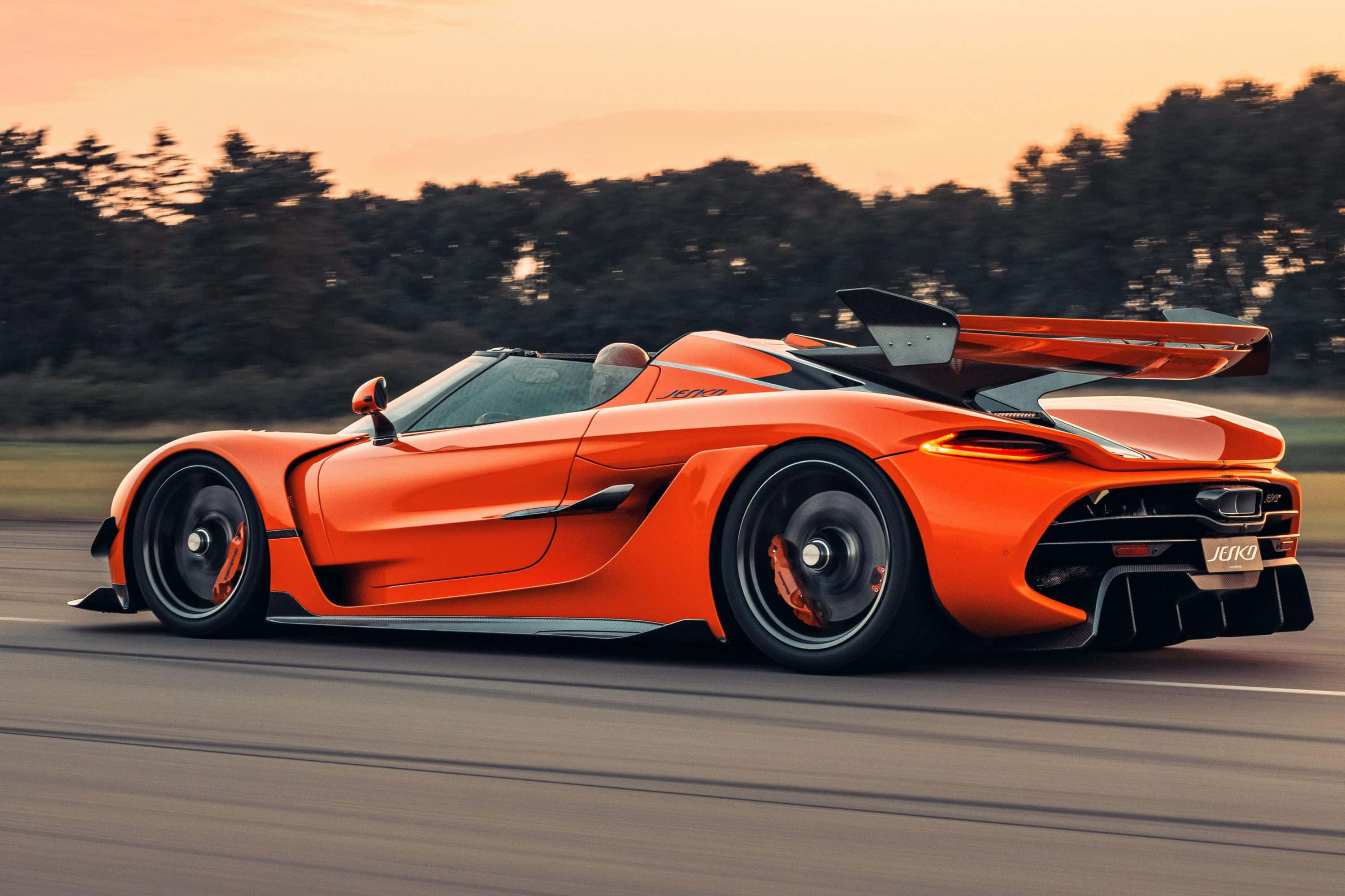 Koenigsegg 桌布 壁紙 — 一輛充滿活力的橙色Koenigsegg Jesko超跑在日落時分沿著道路飛馳，背景是模糊的樹木。