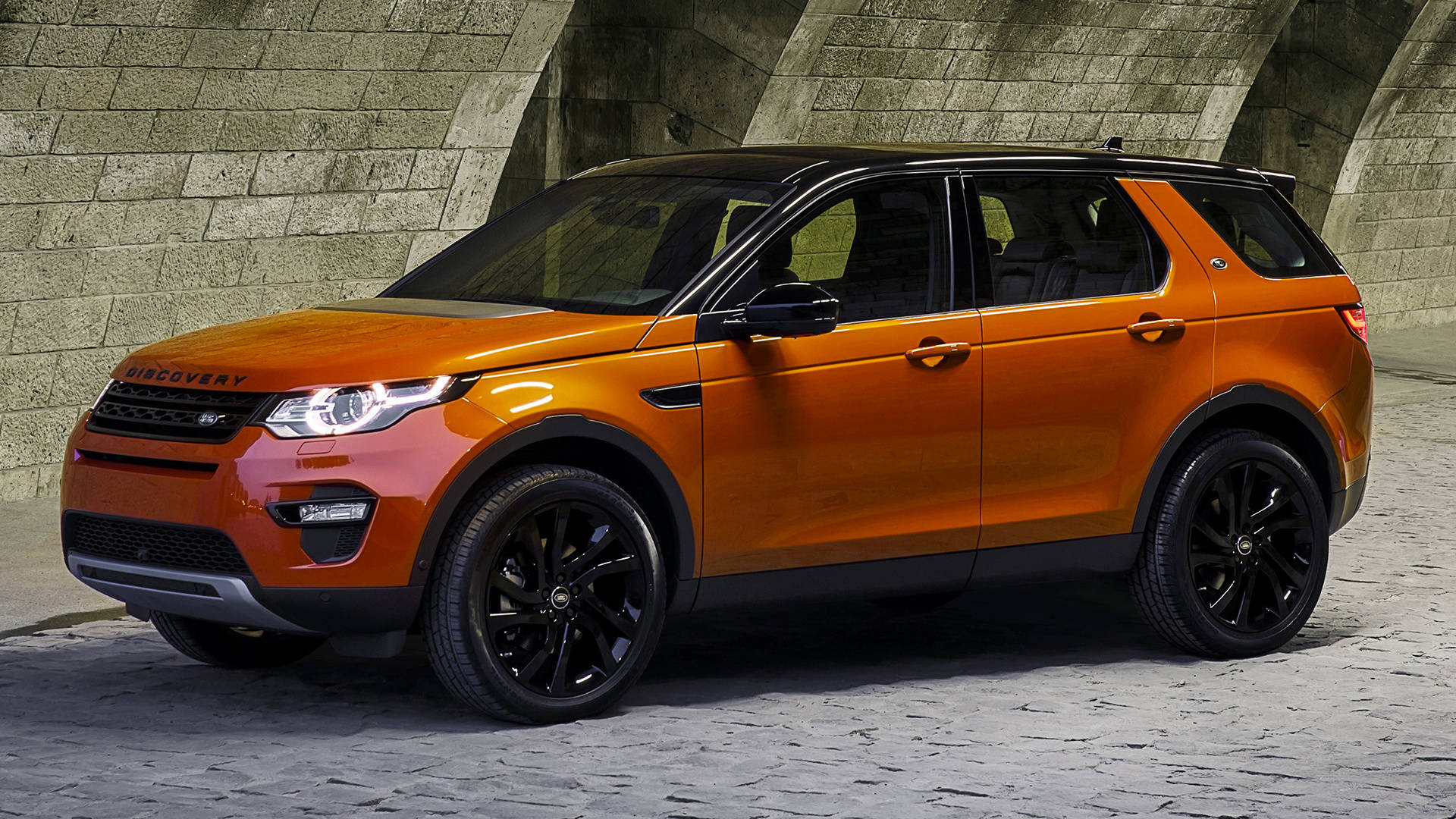 land rover iPhone 桌布 壁紙 — 一輛充滿活力的橙色Land Rover Discovery Sport，帶有黑色飾邊，停放在石拱門下，其時尚設計在明亮的日光下閃閃發光。