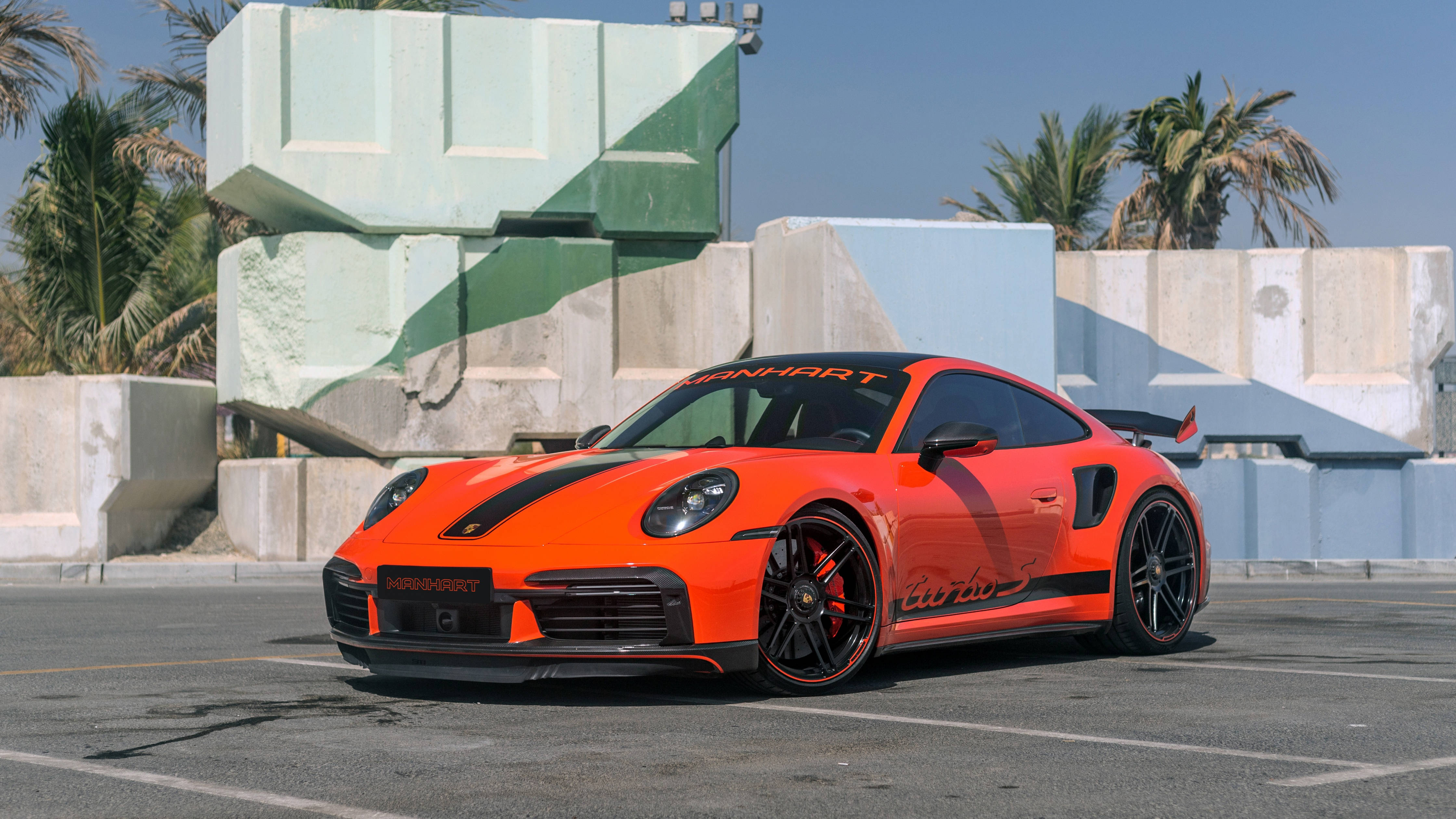 4K 桌布 壁紙 — 一輛充滿活力的橙色Manhart改裝保時捷911 Turbo S停泊在陽光普照的混凝土停車場，背景有棕櫚樹和幾何結構。
