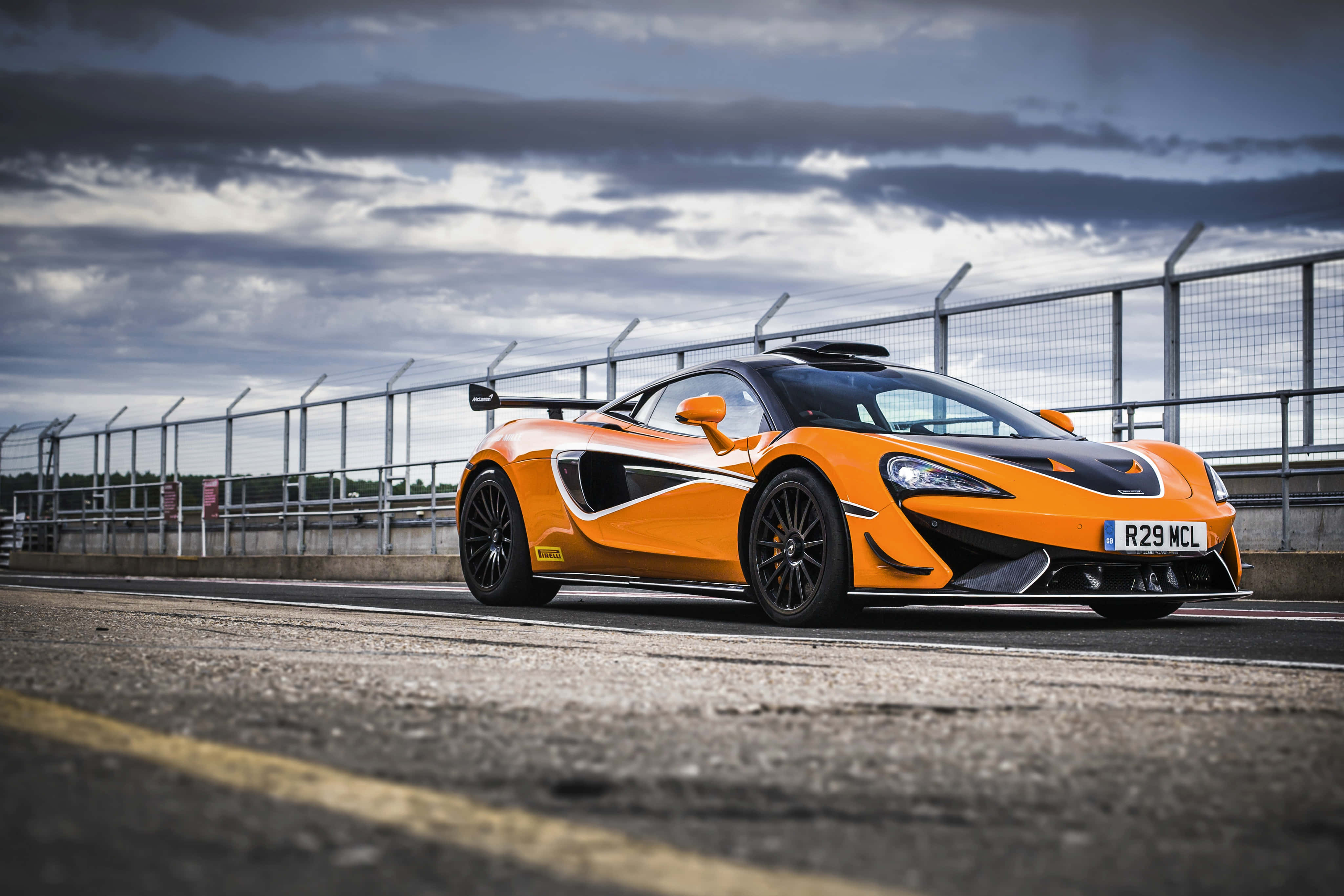 Mclaren 620r 桌布 壁紙 — 一輛充滿活力的橙色McLaren跑車停在賽道上，背景是戲劇性的多雲天空。