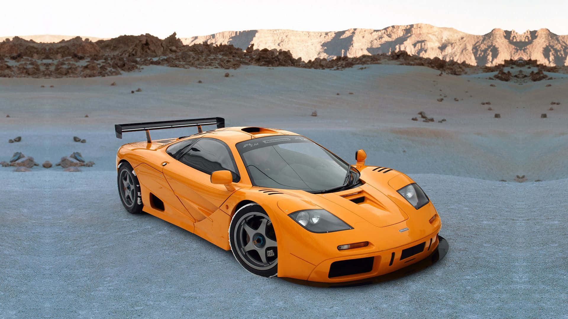 Mclaren F1 桌布 壁紙 — 一輛充滿活力的橙色McLaren F1超級跑車停泊在廣闊的沙漠平原上，背景是岩石山脈，天空明亮。