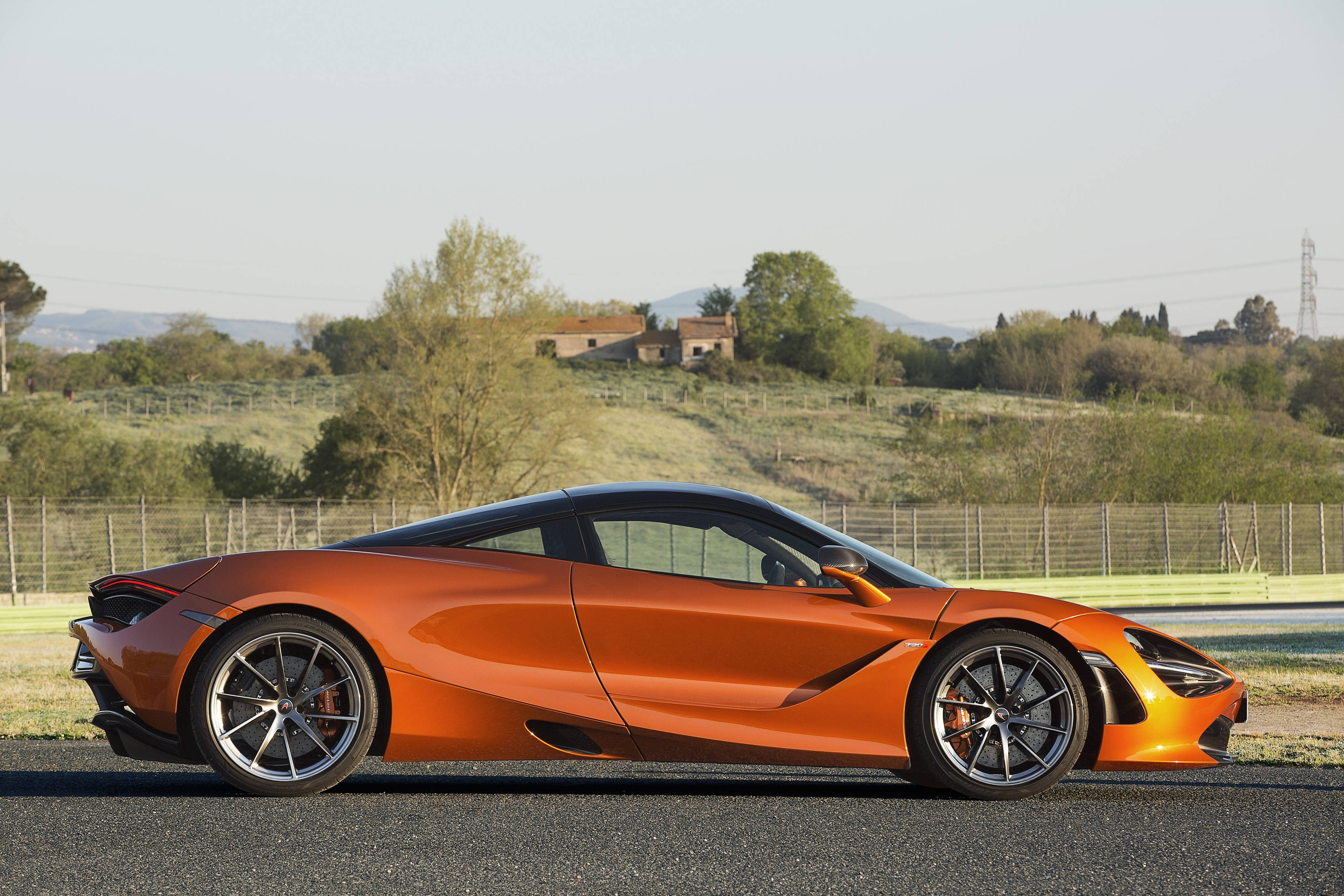 Mclaren 720s 桌布 壁紙 — 一輛充滿活力的橙色McLaren跑車在瀝青賽道上側面閃耀，背景是連綿的綠色山丘和遠處的建築物，天空朦朧。