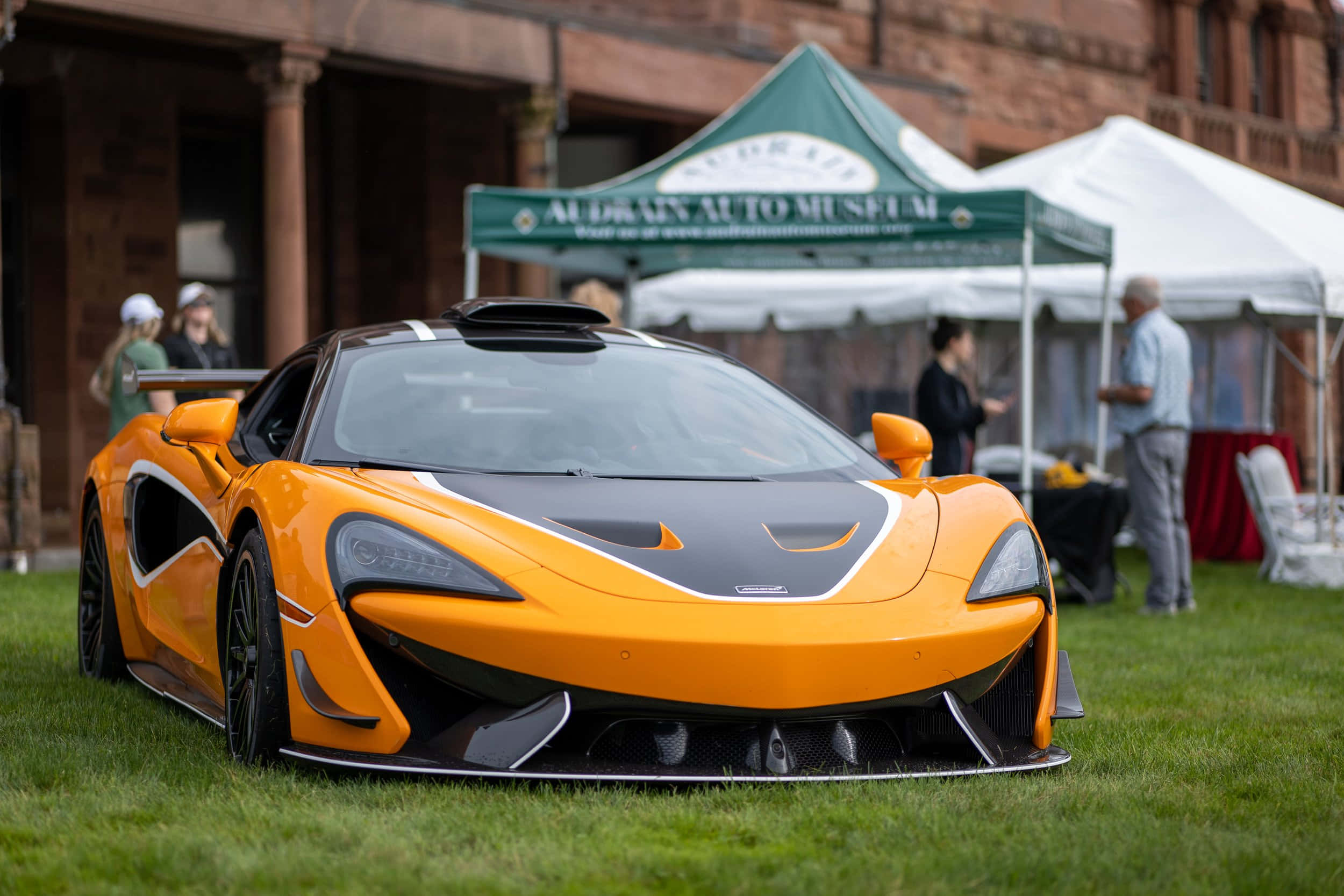 Mclaren 620r 桌布 壁紙 — 一輛充滿活力的橙色McLaren跑車帶有黑色飾邊，突出地停在綠色草地上，背景是戶外汽車活動，有人群和帳篷。