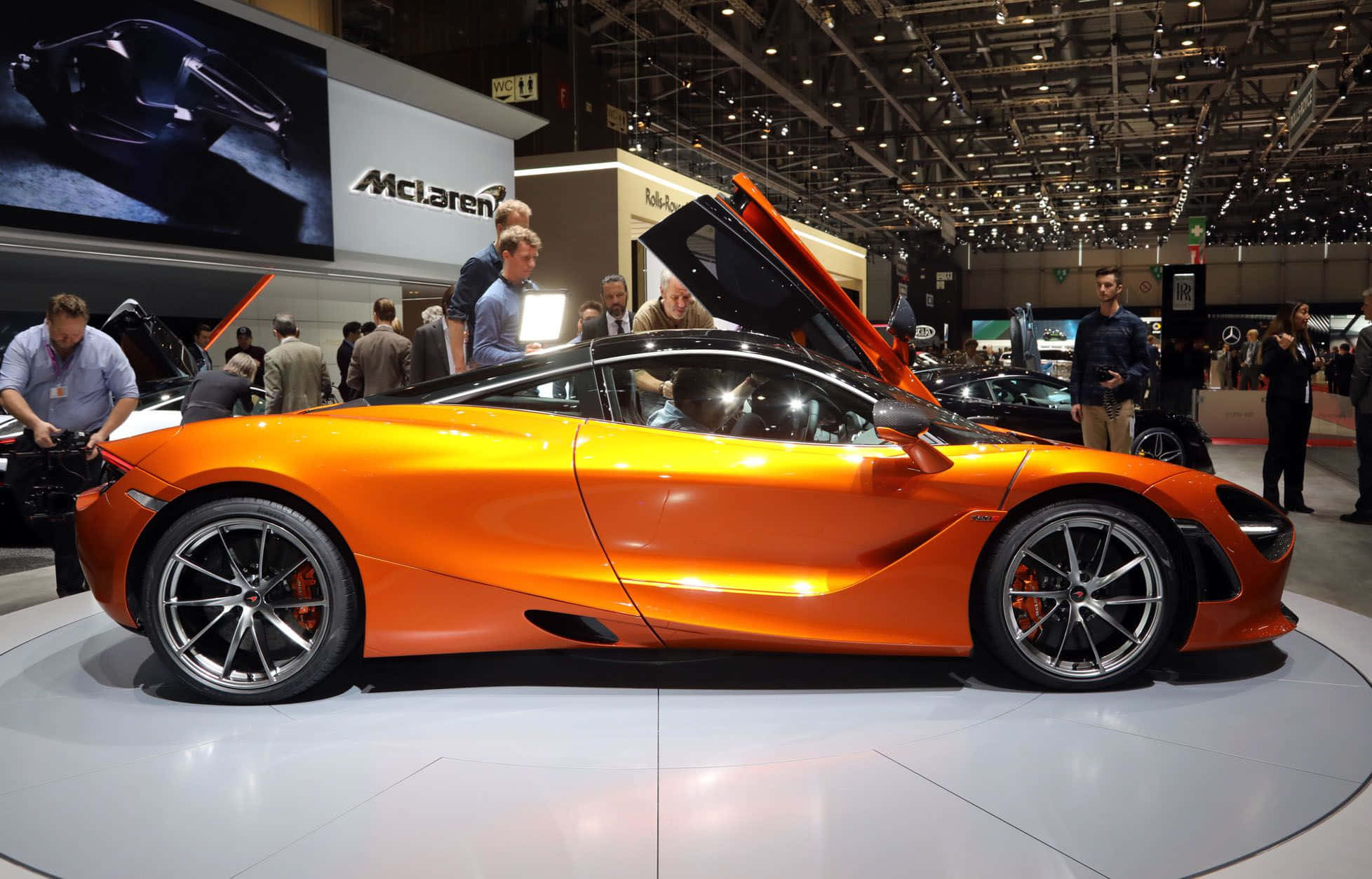show 桌布 壁紙 — 一輛充滿活力的橙色McLaren超級跑車，蝴蝶門打開，在繁忙的車展的圓形平台上展示。