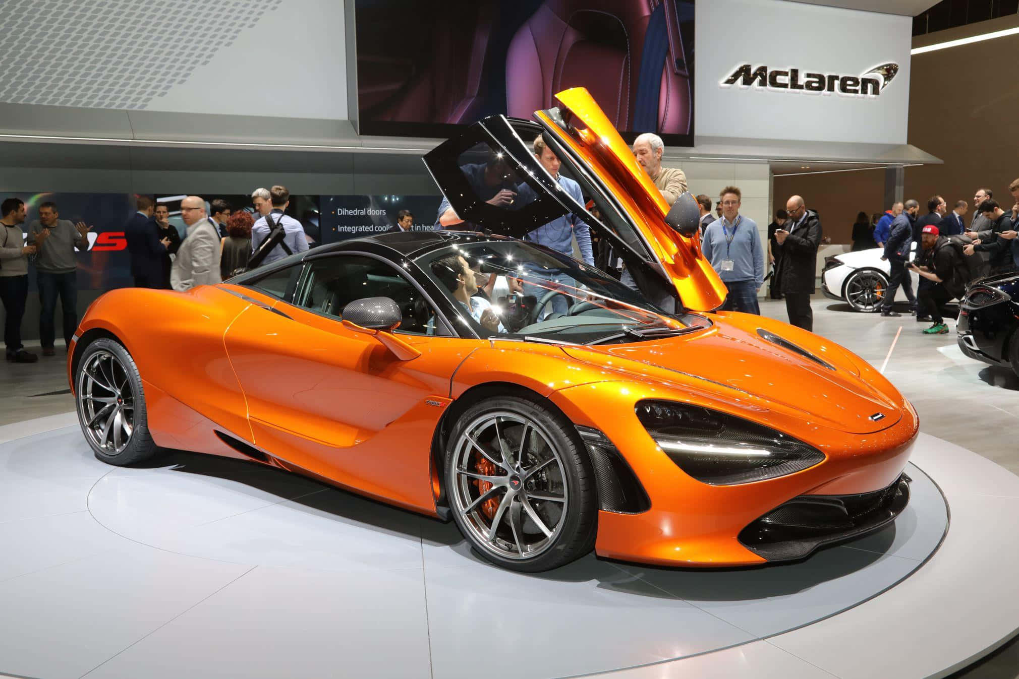 Mclaren 720s 桌布 壁紙 — 一輛充滿活力的橙色McLaren跑車，鷗翼門打開，在車展上展出，周圍是欣賞的人群。