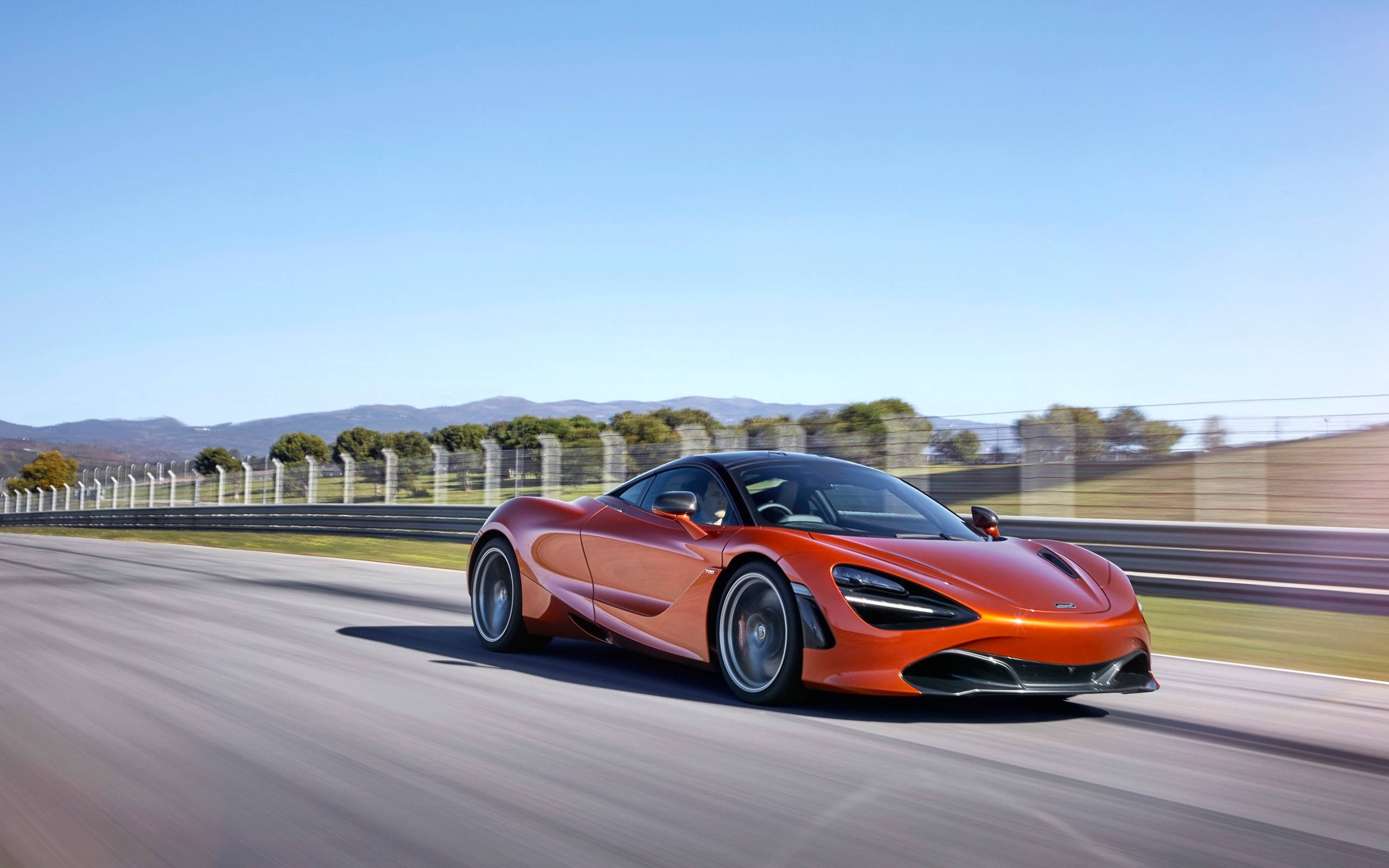 mclaren 720s 4K 桌布 壁紙 — 一輛充滿活力的橙色McLaren跑車在晴朗的藍天下，沿著賽道高速行駛，呈現動態模糊效果。