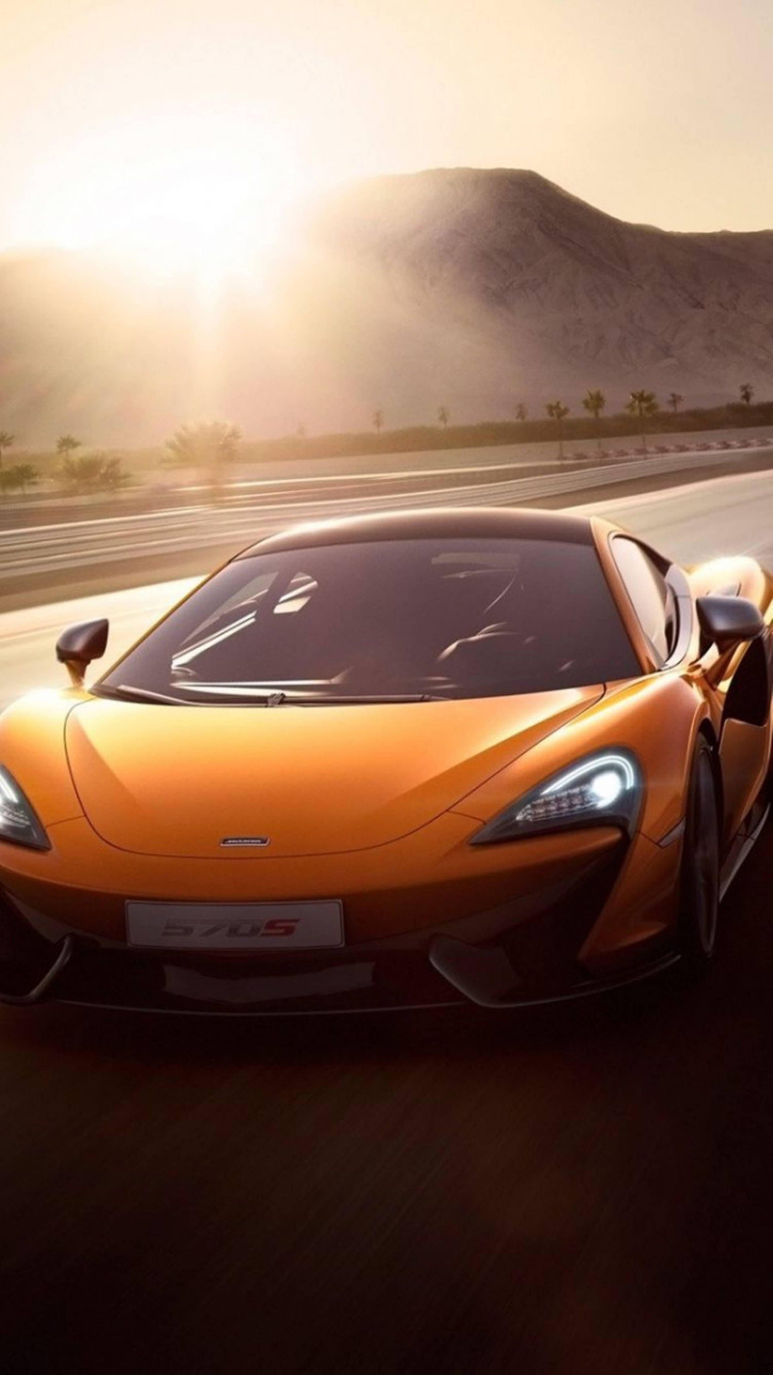 Luxury 桌布 壁紙 — 一輛充滿活力的橙色McLaren 570S在日出時沿著沙漠高速公路奔馳，背景有山脈。