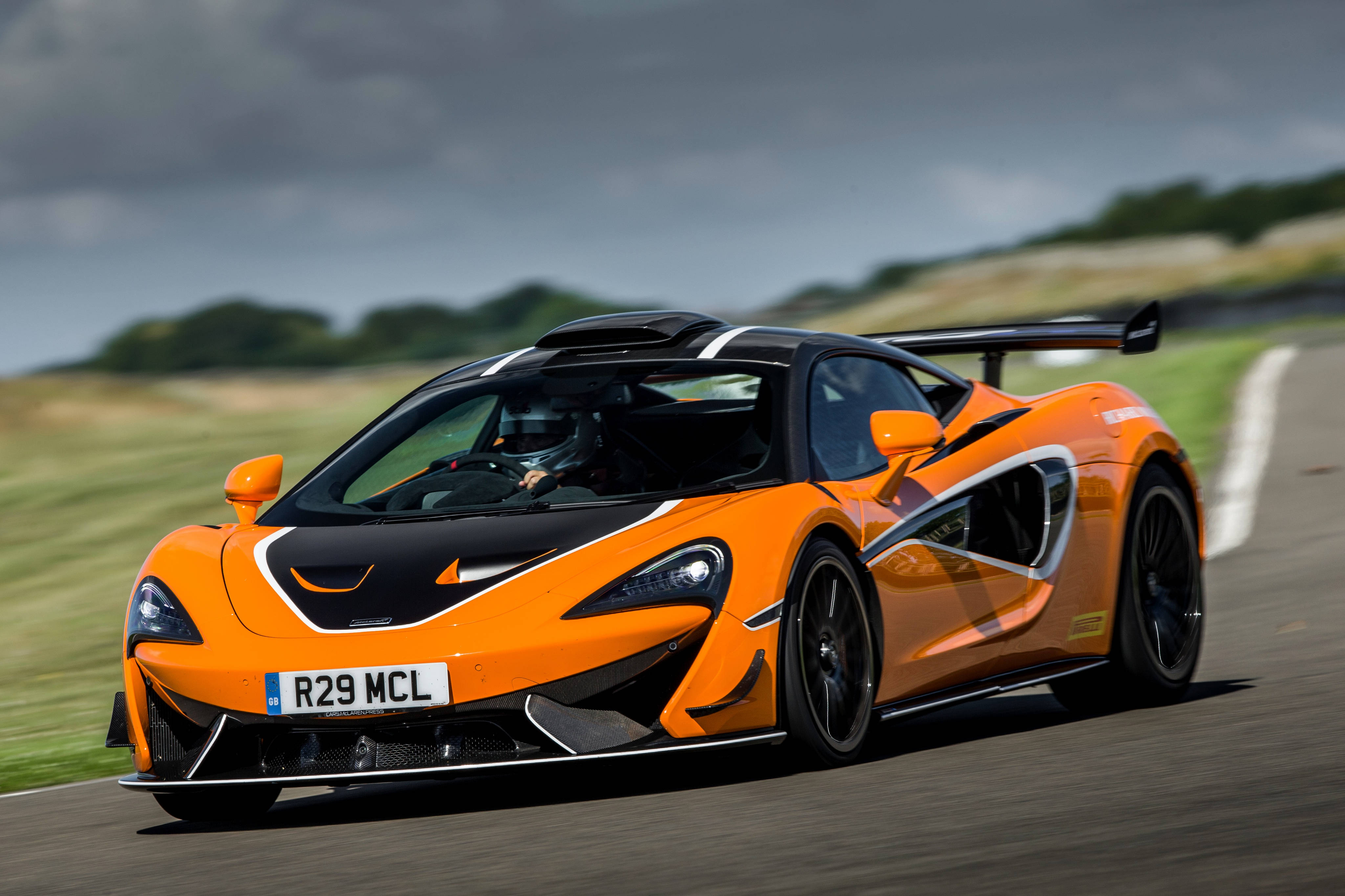 Auto Racing 桌布 壁紙 — 一輛充滿活力的橙色McLaren跑車在賽道上疾馳，背景是模糊的綠色山丘和陰天。