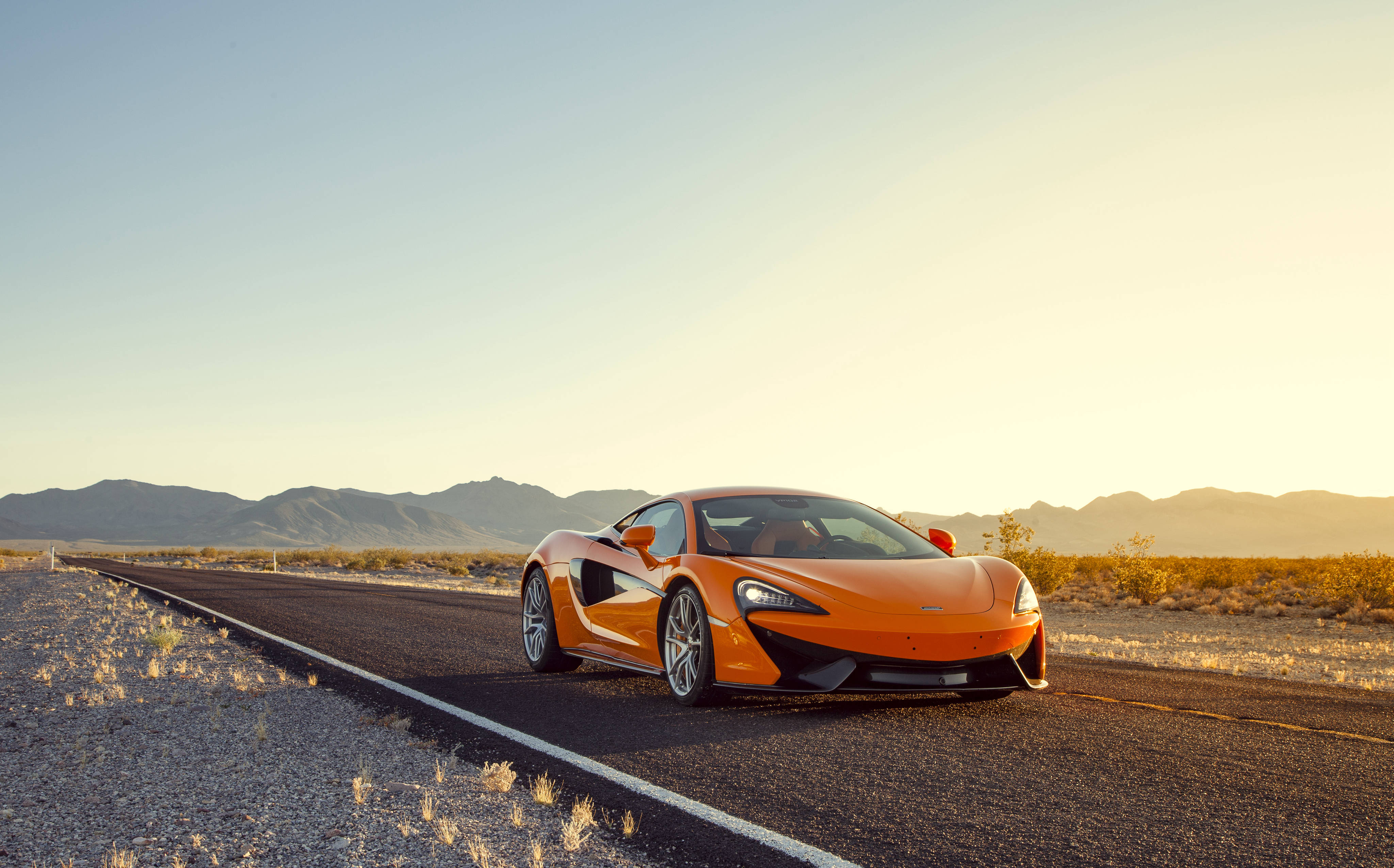 麥拉倫 桌布 壁紙 — 一輛充滿活力的橙色McLaren跑車在黃金時段沿沙漠高速公路行駛，遠山在柔和天空下。