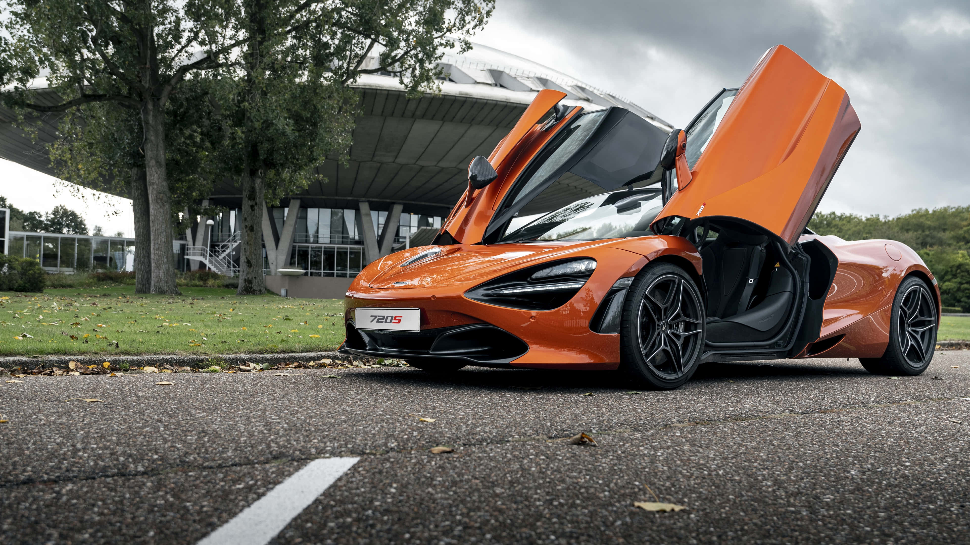 4K Mclaren 720s 桌布 壁紙 — 一輛充滿活力的橙色McLaren 780S超級跑車，鷗翼車門打開，停泊在瀝青路上，附近有樹木和現代建築，天空多雲。