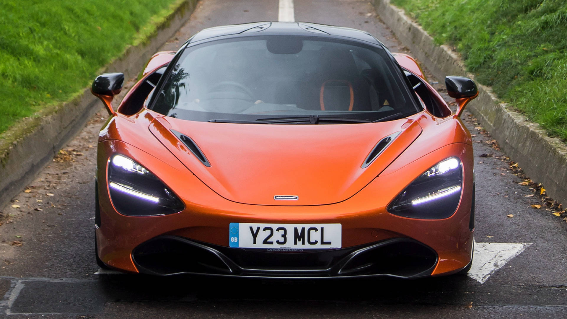 mclaren 720s 4K 桌布 壁紙 — 一輛充滿活力的橙色McLaren 720S跑車在濕滑的道路上向前行駛，兩旁是綠草和落葉。