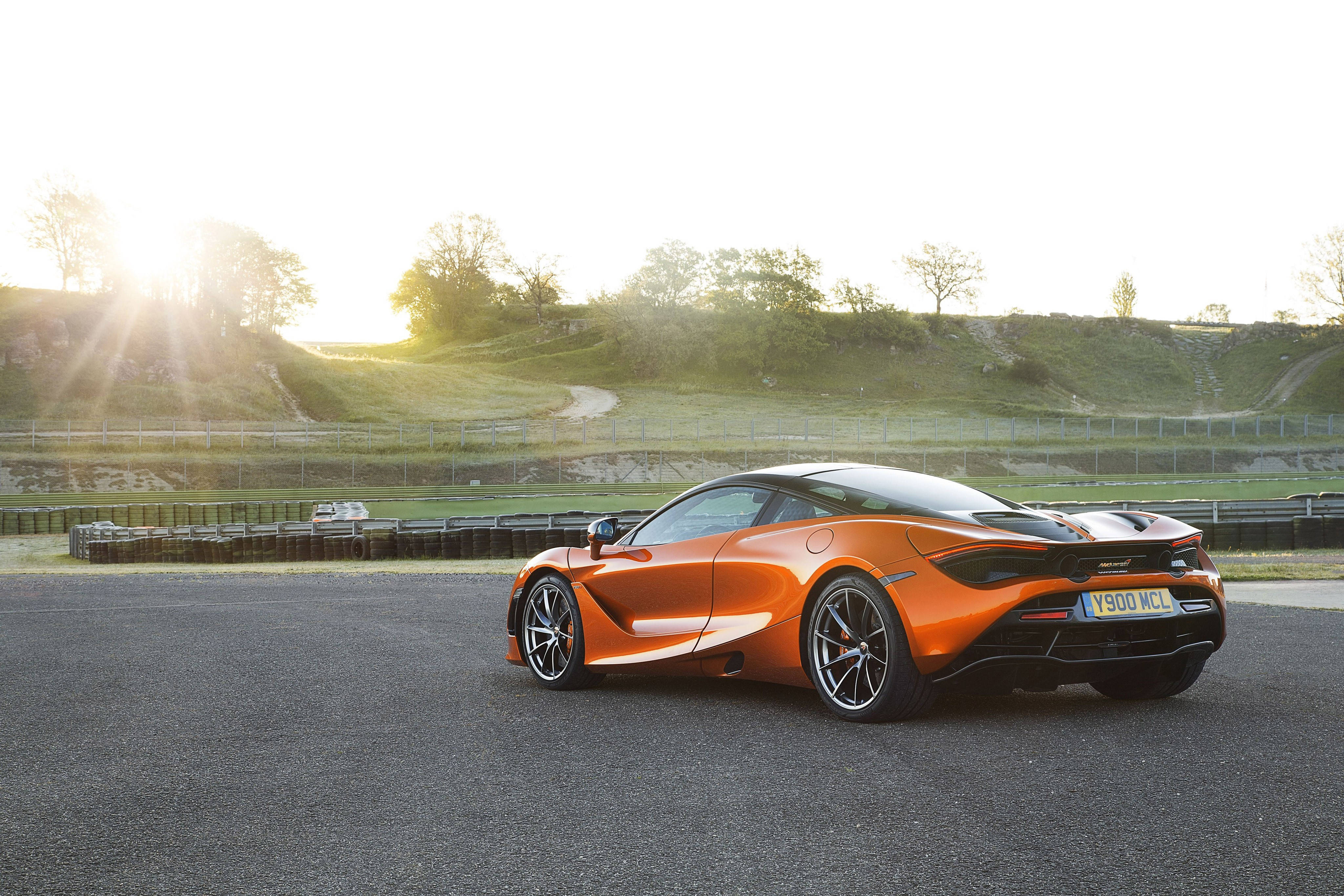Mclaren 720s 桌布 壁紙 — 一輛充滿活力的橙色McLaren跑車在瀝青賽道上閃閃發光，金色陽光穿過樹木灑在起伏的綠色山坡上。