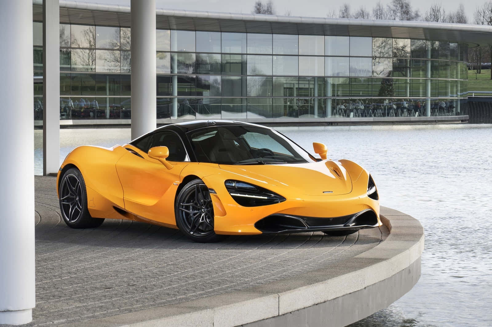 汽車 桌布 壁紙 — 一輛充滿活力的橙色McLaren 720S跑車停泊在平靜水邊的弧形石台上，背景是一座現代玻璃建築。