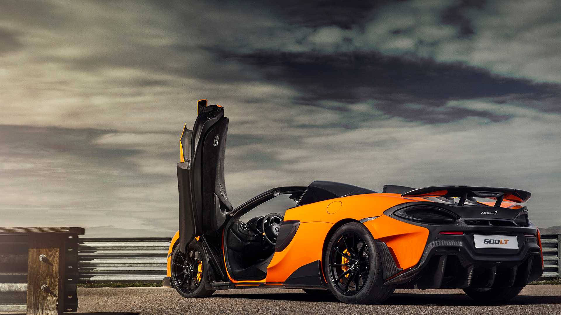 跑車 桌布 壁紙 — 一輛充滿活力的橙色McLaren 600LT跑車，剪刀門打開，停在賽道上，背景是戲劇性的多雲天空。