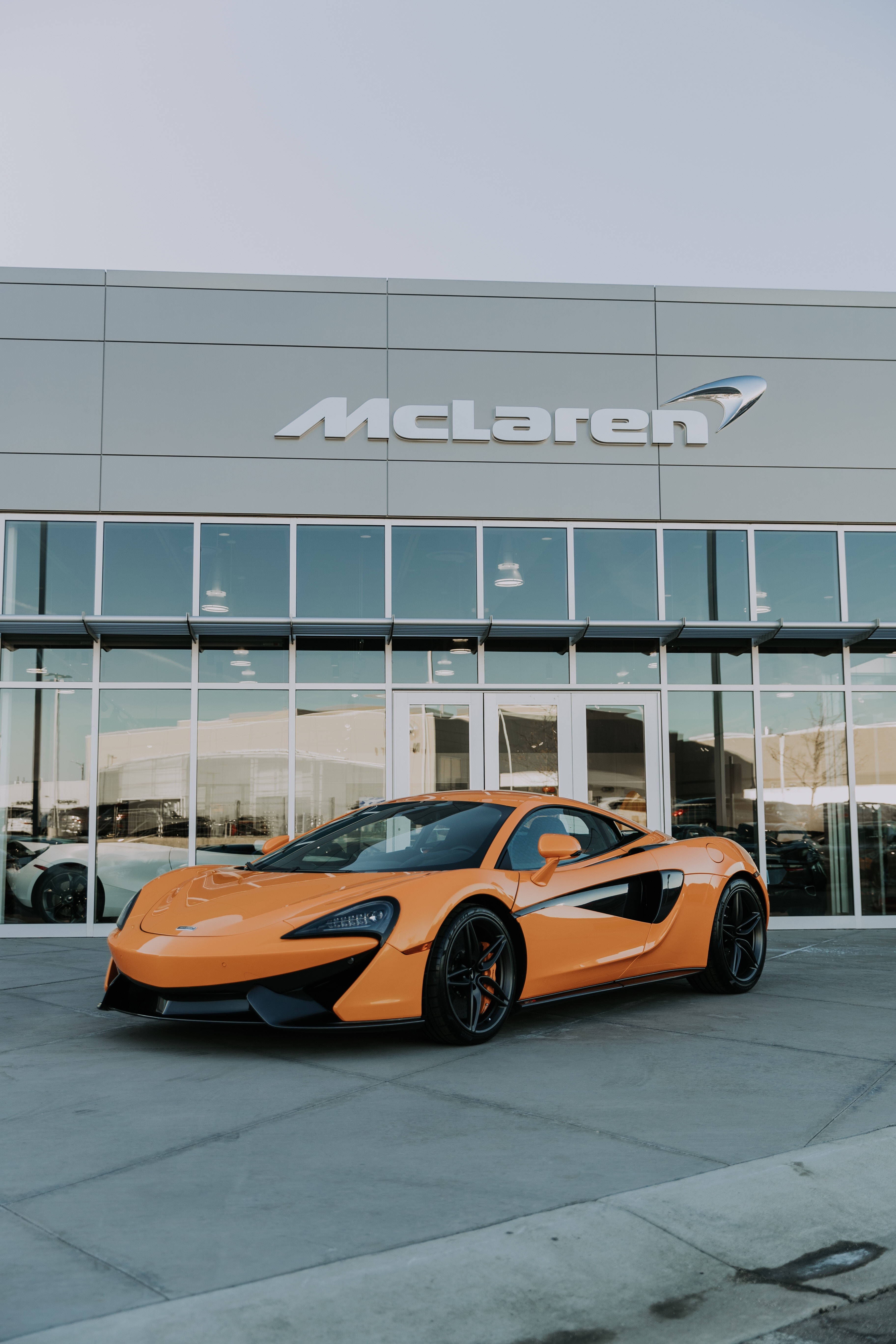 跑車 桌布 壁紙 — 一輛充滿活力的橙色McLaren跑車停在一間現代化展示間前，該展示間設有玻璃窗和灰色外牆。