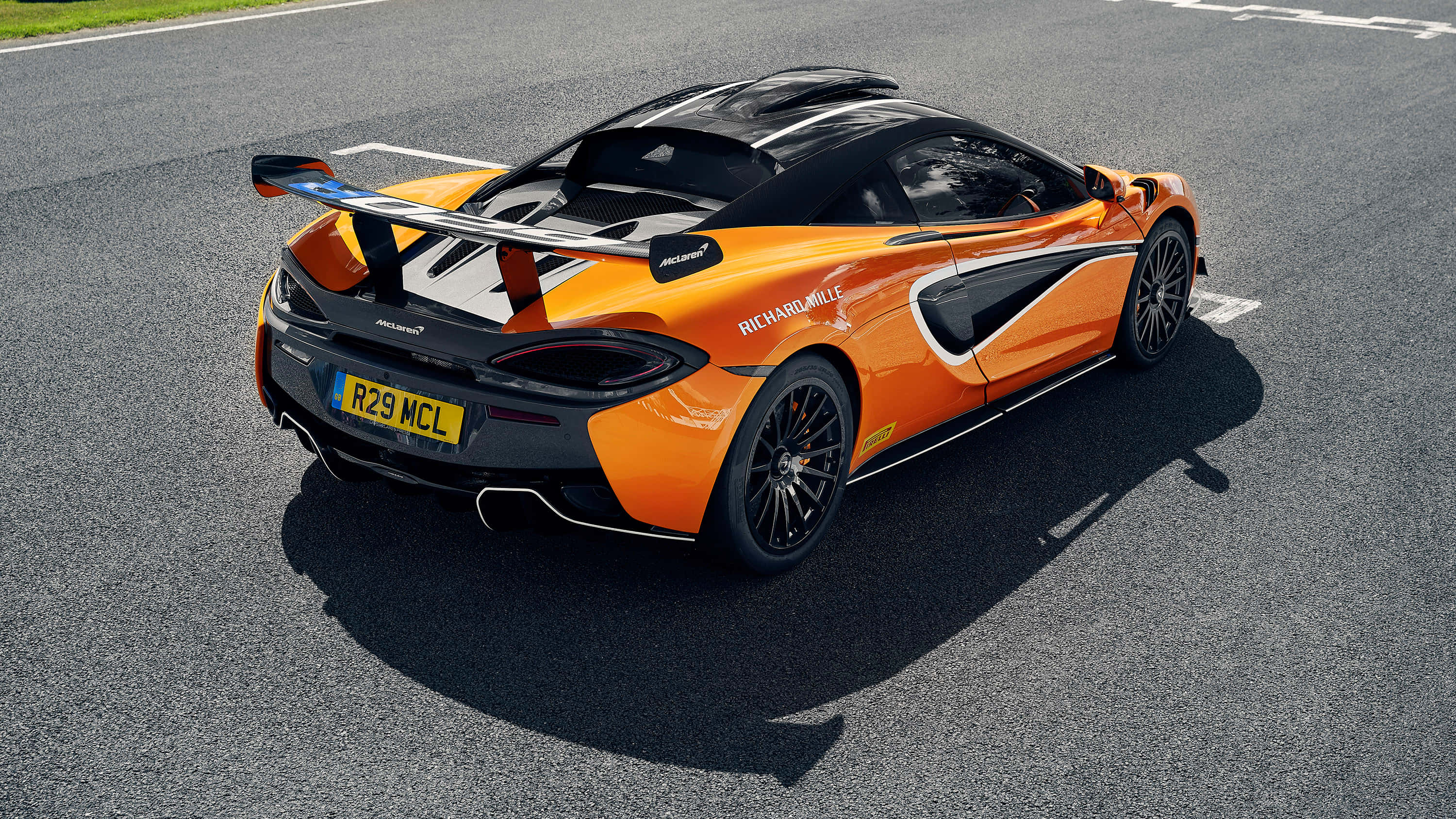 Mclaren 620r 桌布 壁紙 — 一輛充滿活力的橙色McLaren跑車，帶有黑色飾件和大型尾翼，停在陽光普照的賽道上。