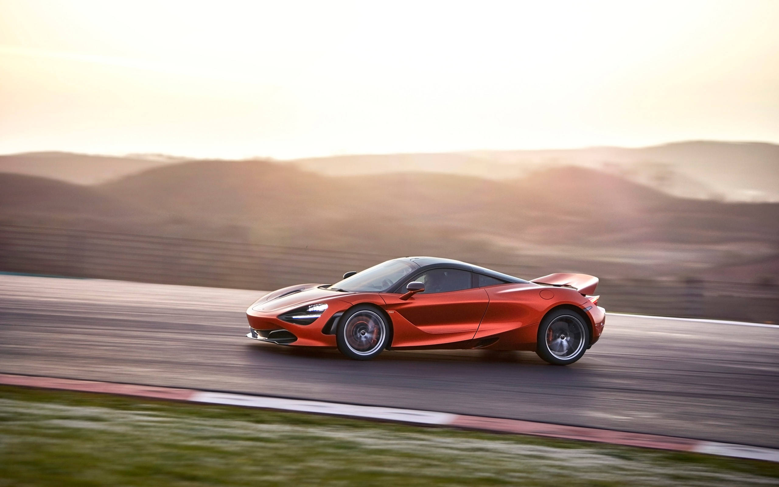 mclaren 720s 4K 桌布 壁紙 — 一輛充滿活力的橙色McLaren 720S在日落時分沿著賽道飛馳，背景是模糊的山丘。