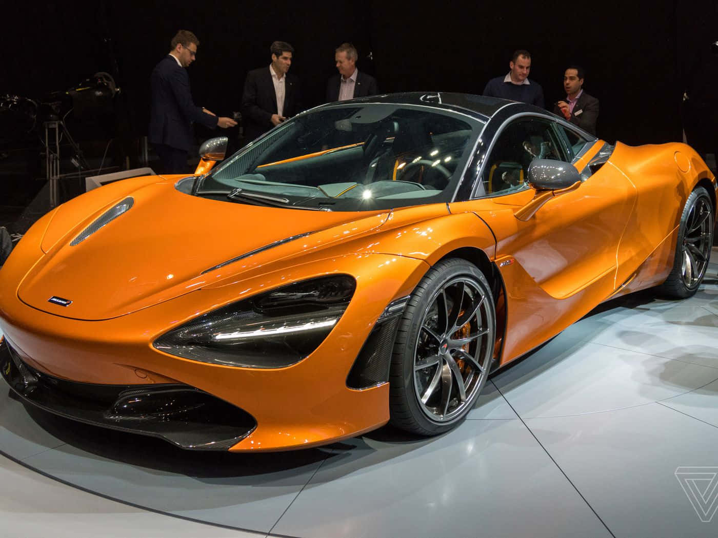 汽車 桌布 壁紙 — 一輛充滿活力的橙色McLaren 720S超級跑車在車展的明亮燈光下閃閃發光，身穿西裝的男士們正在觀看。