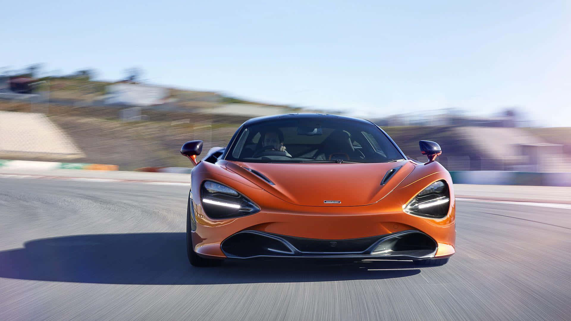 麥拉倫 720S 桌布 壁紙 — 一輛充滿活力的橙色McLaren跑車在賽道上疾馳，動態模糊傳達出明亮日光下的高速感。