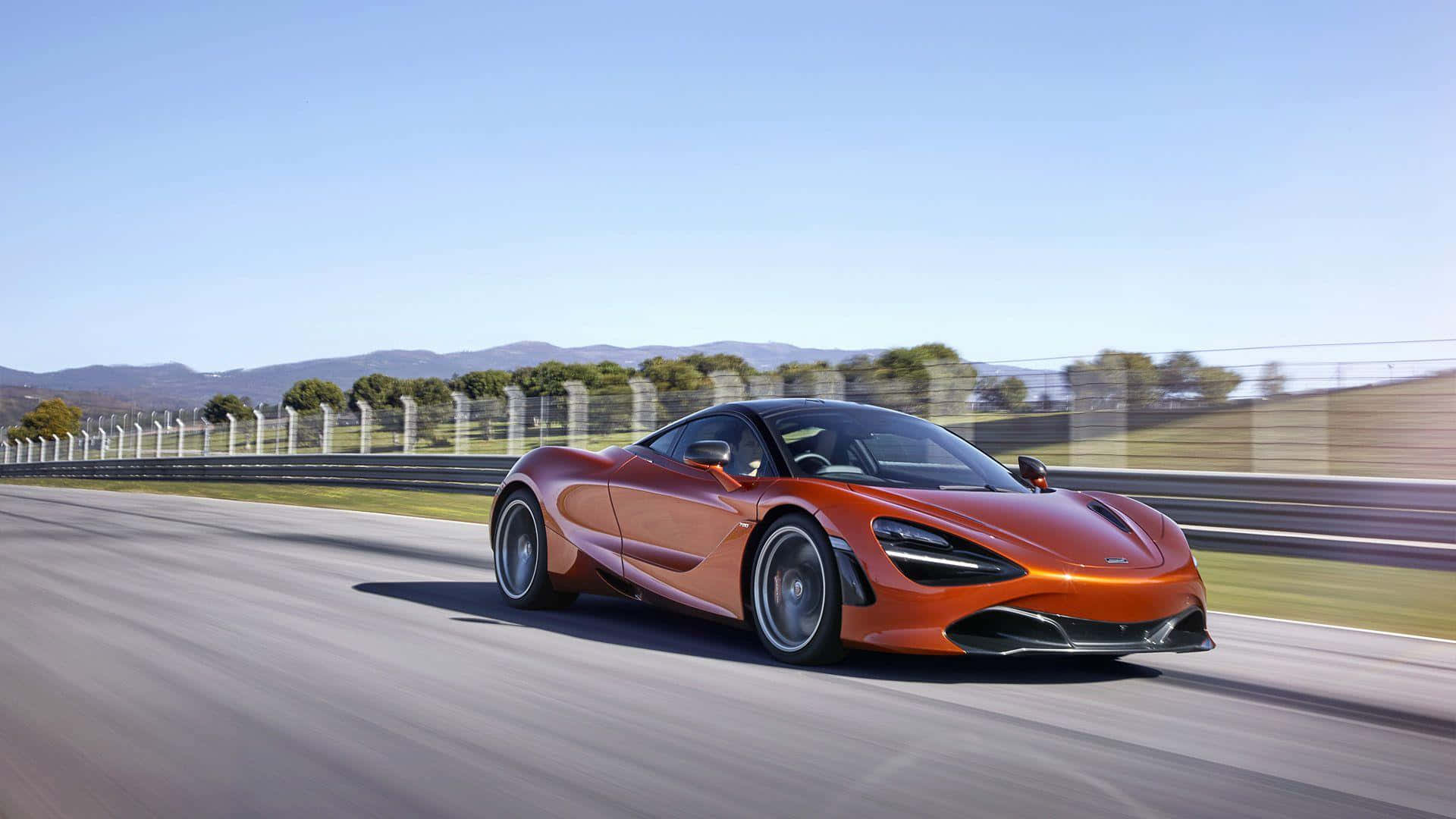 Mclaren 720s 桌布 壁紙 — 一輛充滿活力的橙色McLaren 720S在陽光明媚的賽道上疾馳，背景是模糊的動感和遠處的山丘，天空清澈湛藍。