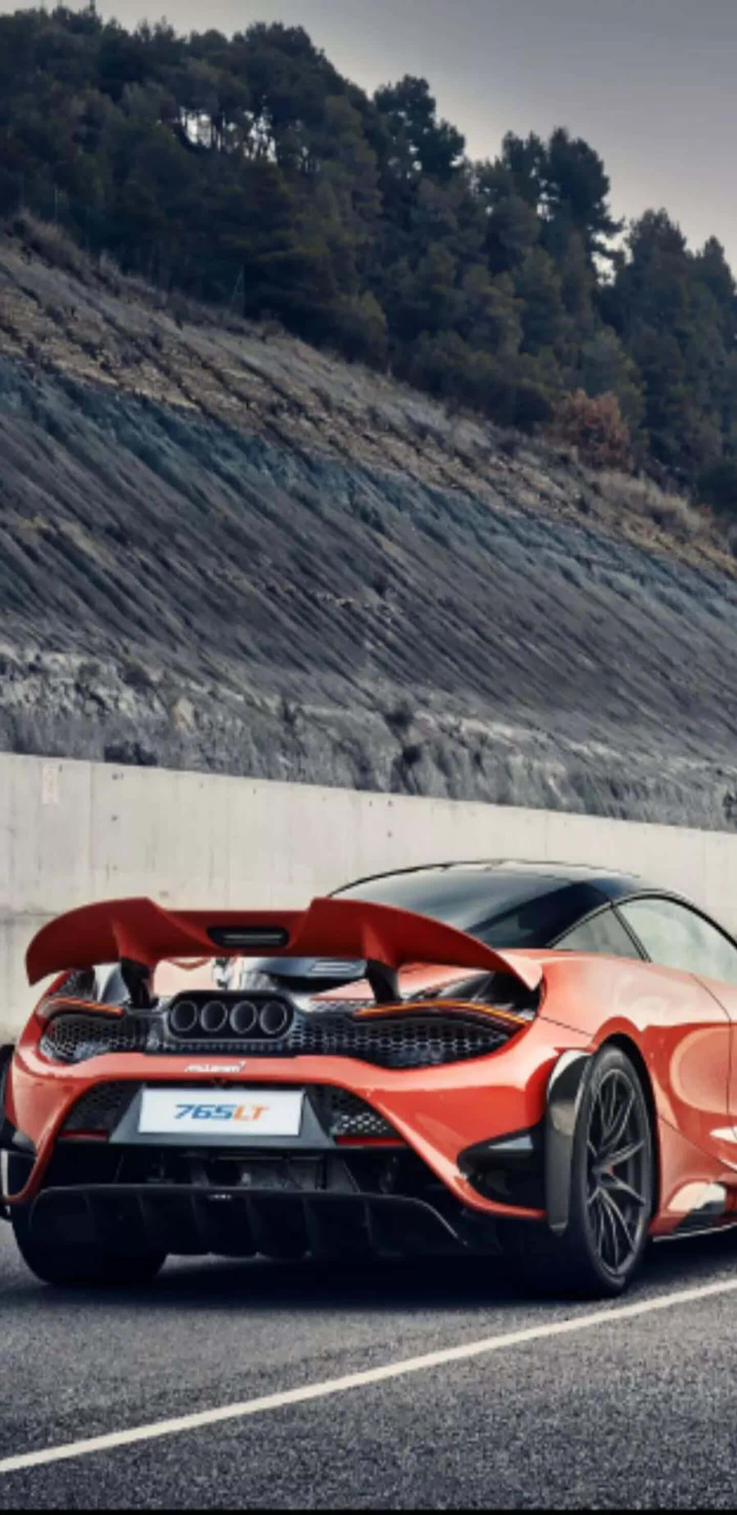 Pixel 3xl 桌布 壁紙 — 一輛充滿活力的橙色McLaren 765LT超級跑車，尾翼升起，停在柏油路上，旁邊是混凝土護欄和佈滿樹木的岩石山坡。
