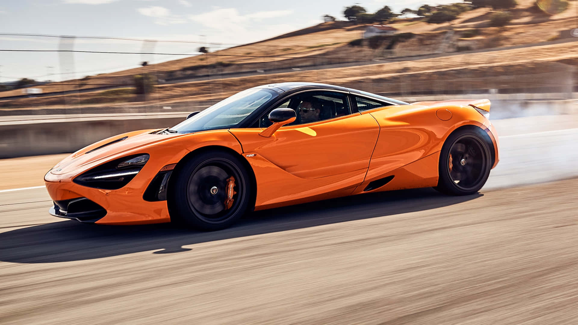 Race Track 桌布 壁紙 — 一輛充滿活力的橙色McLaren 720S在陽光明媚的賽道上疾馳，動態模糊效果凸顯其強勁動力和流線型空氣動力學設計。