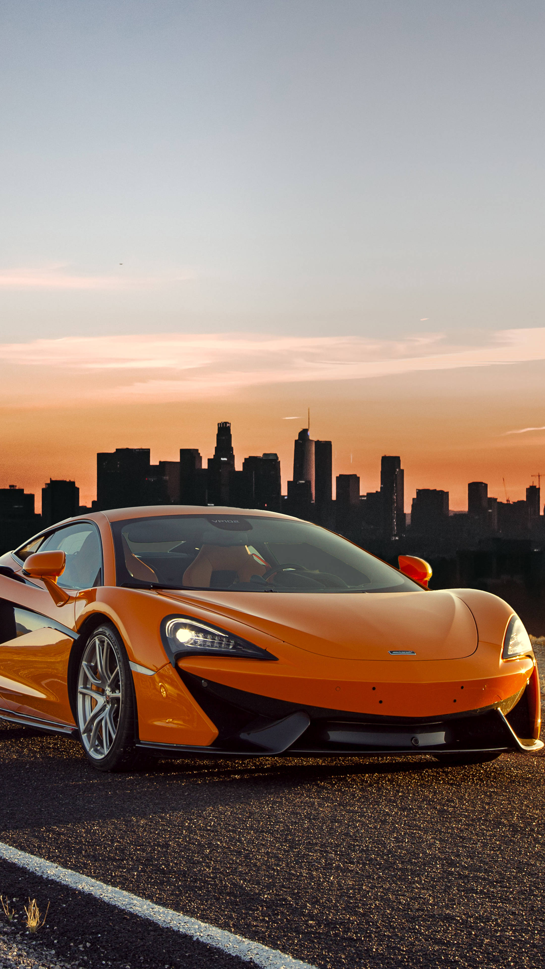 橘色 桌布 壁紙 — 一輛充滿活力的橙色McLaren跑車停泊在道路上，背景是日落時分的城市天際線。
