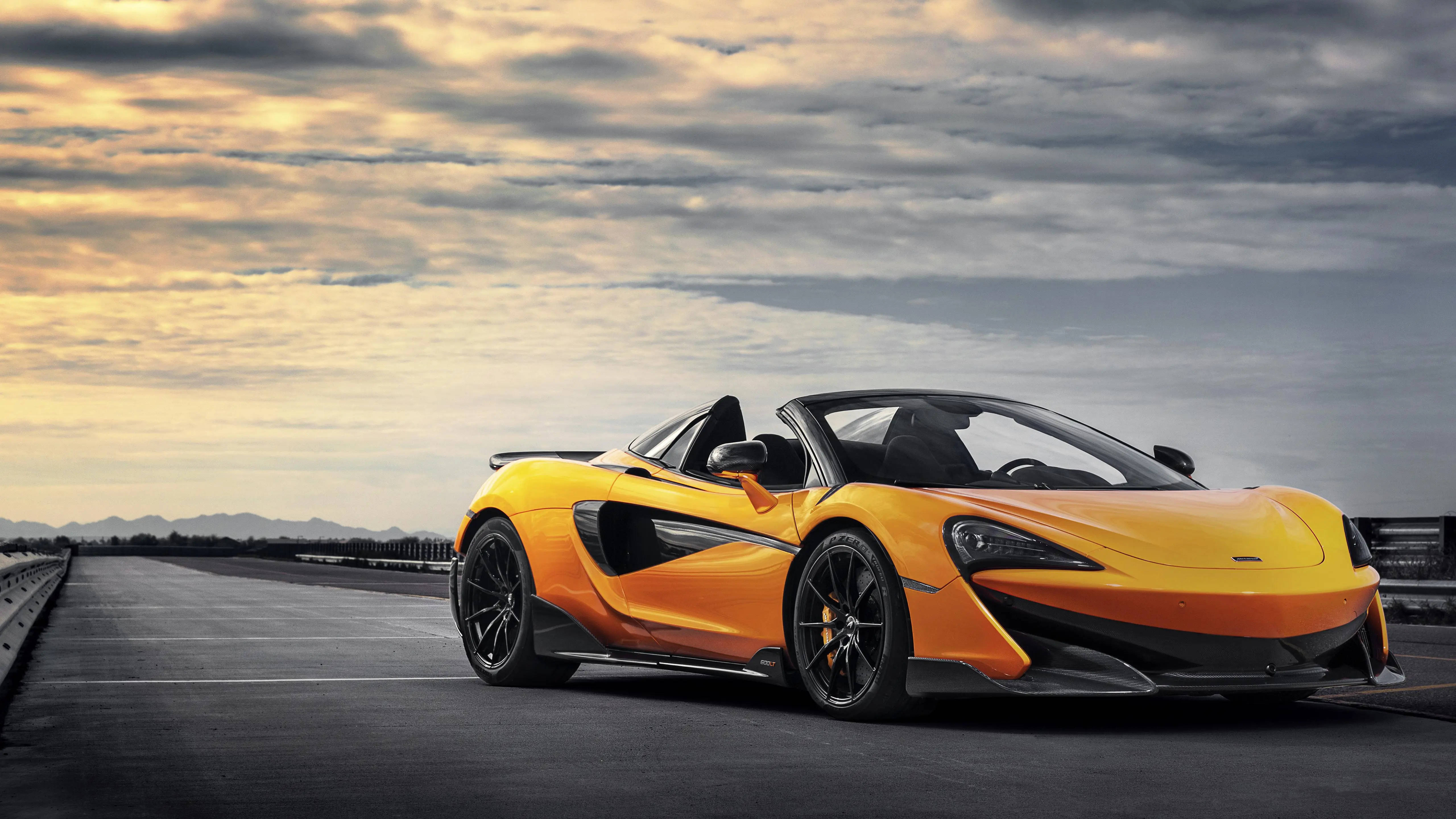 跑車 桌布 壁紙 — 一輛充滿活力的橙色McLaren跑車，帶有黑色點綴，停泊在賽道上，背景是日落時分戲劇性的多雲天空。