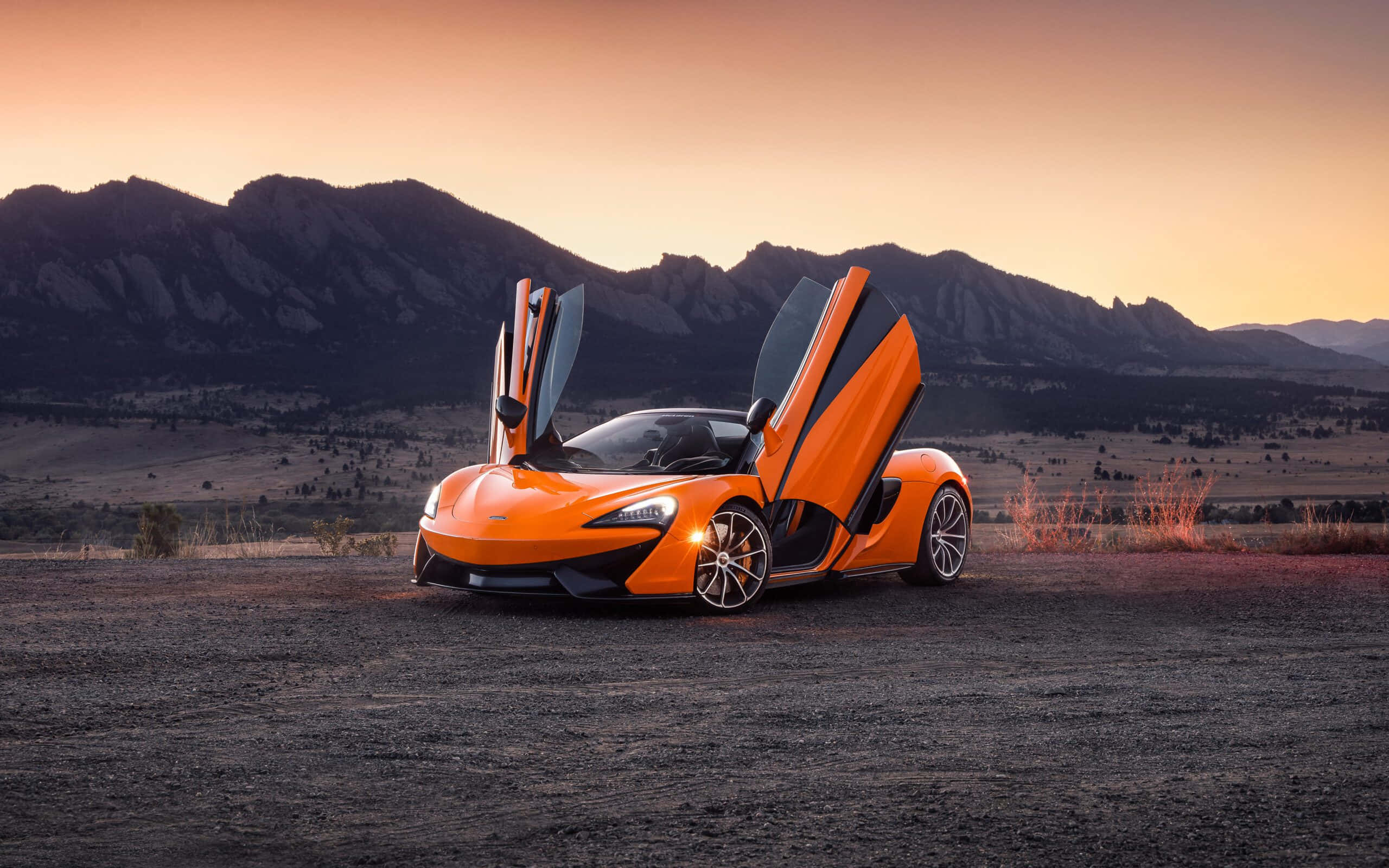 麥拉倫 桌布 壁紙 — 一輛充滿活力的橙色McLaren跑車，鷗翼車門打開，停在夕陽下的礫石平原上，背景是崎嶇的山脈。