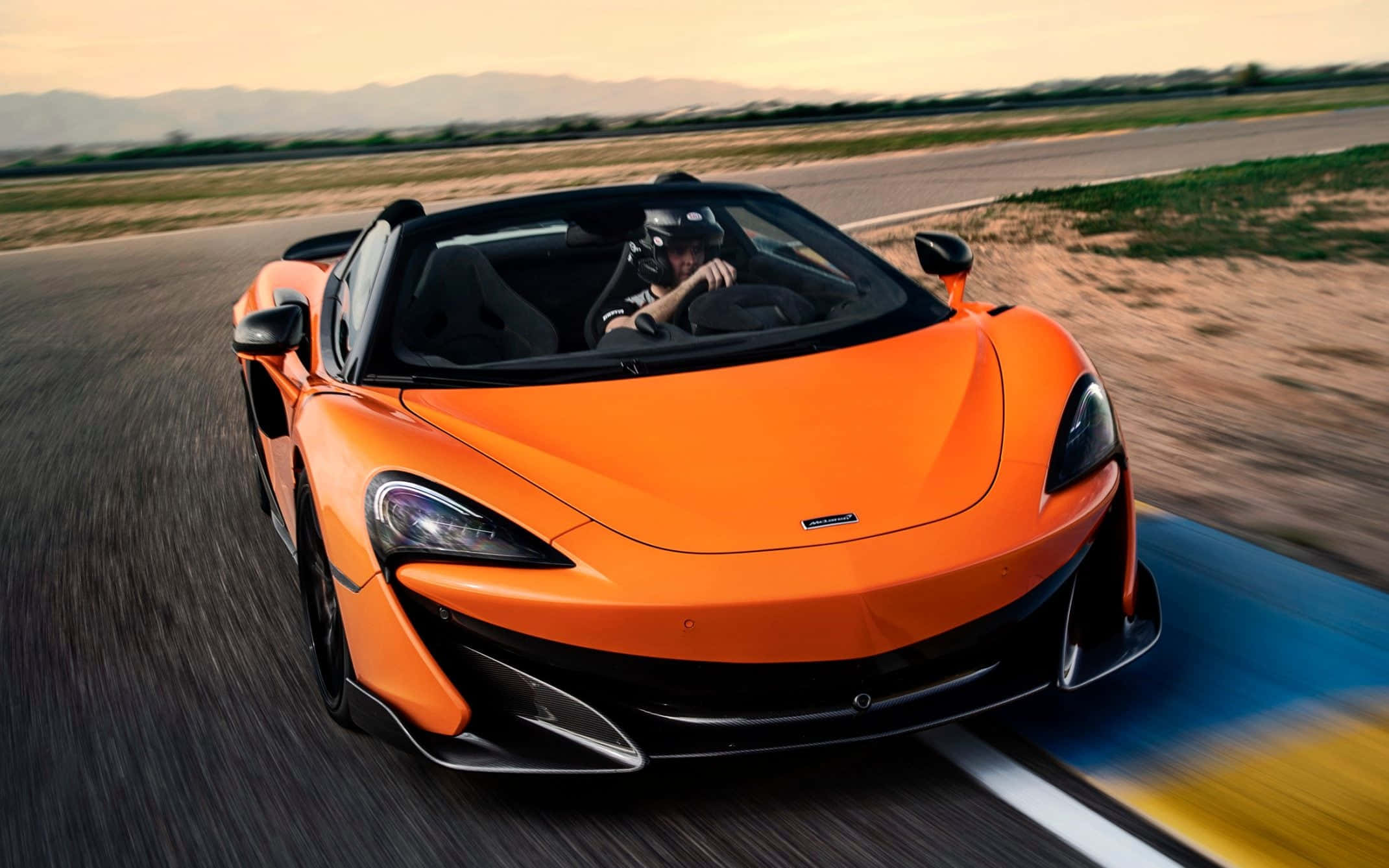 麥拉倫 桌布 壁紙 — 一輛充滿活力的橙色McLaren 570S敞篷跑車在日落時分沿著沙漠賽道疾馳，帶有模糊的動感和遠山。