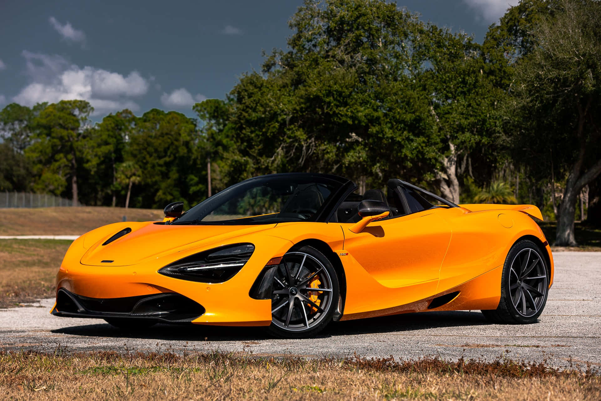 草地 桌布 壁紙 — 一輛充滿活力的橙色McLaren 720S Spider敞篷車停在碎石地上，周圍有茂密的綠樹，天空局部多雲。