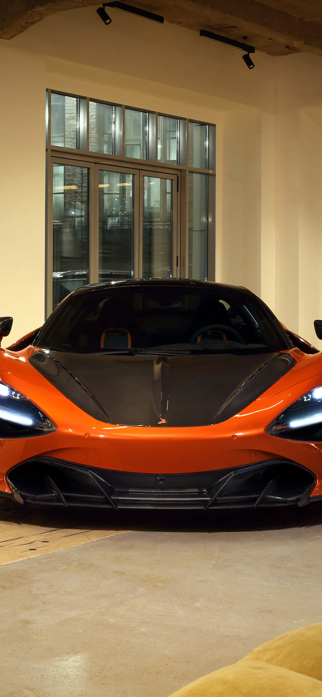 iPhone Xs Mclaren 720s 桌布 壁紙 — 一輛充滿活力的橙色McLaren跑車，帶有黑色飾邊，在室內聚光燈下靠近大窗戶展示。
