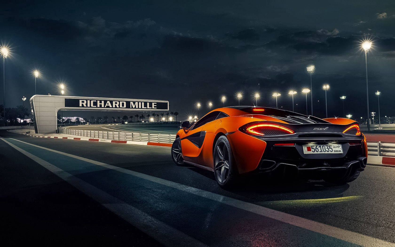 橘色 桌布 壁紙 — 一輛充滿活力的橙色McLaren跑車在夜間沿著賽道滑行，頭頂上方有明亮的泛光燈和一個「RICHARD MILLE」拱門。