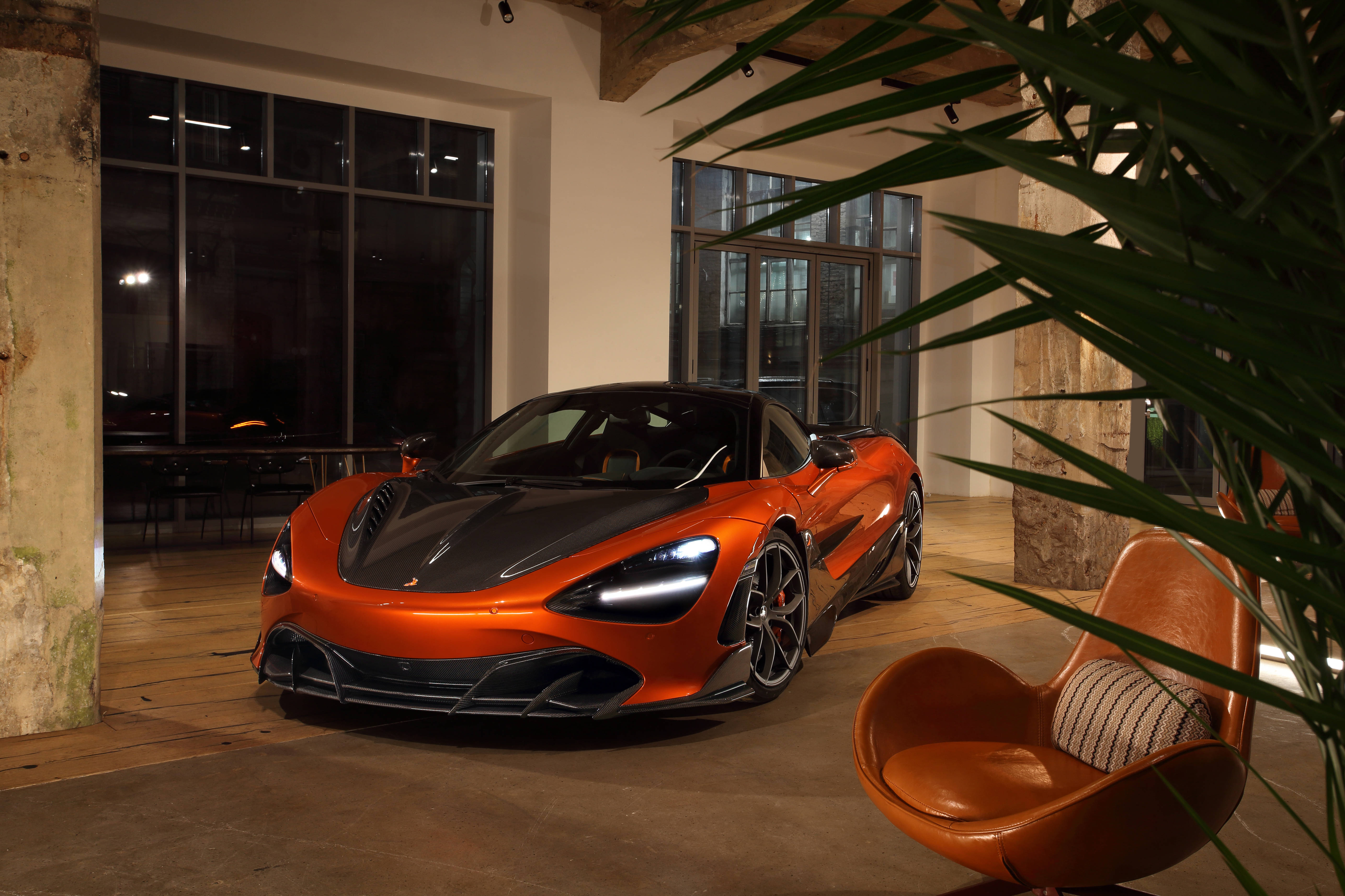 麥拉倫 720S 桌布 壁紙 — 一輛充滿活力的橙色McLaren跑車在溫暖的室內燈光下，在皮革椅和熱帶植物旁邊閃閃發光。