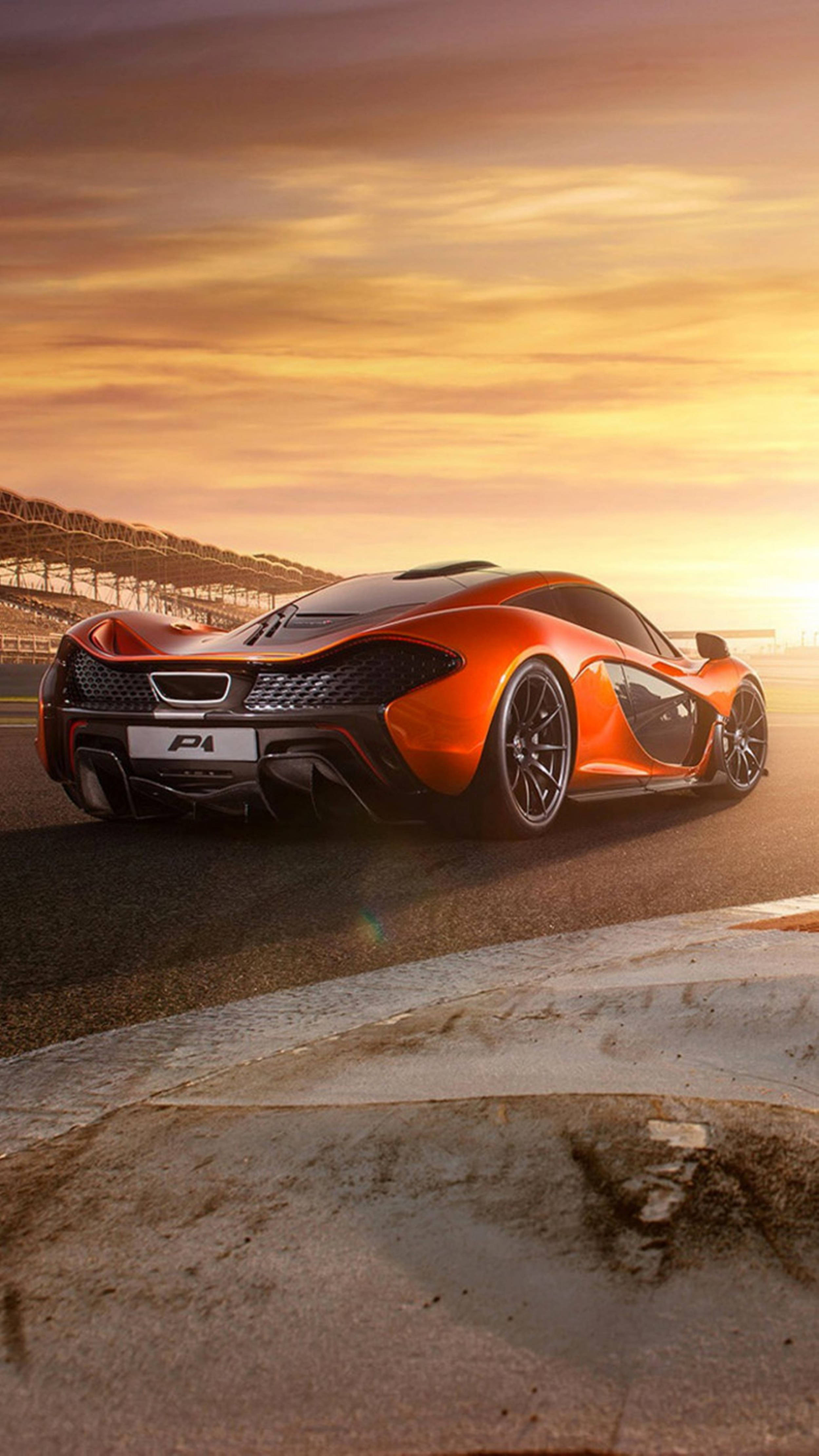 日落 桌布 壁紙 — 一輛充滿活力的橙色McLaren P1超級跑車停在賽道上，背景是金色日落、戲劇性的天空和混凝土護欄。