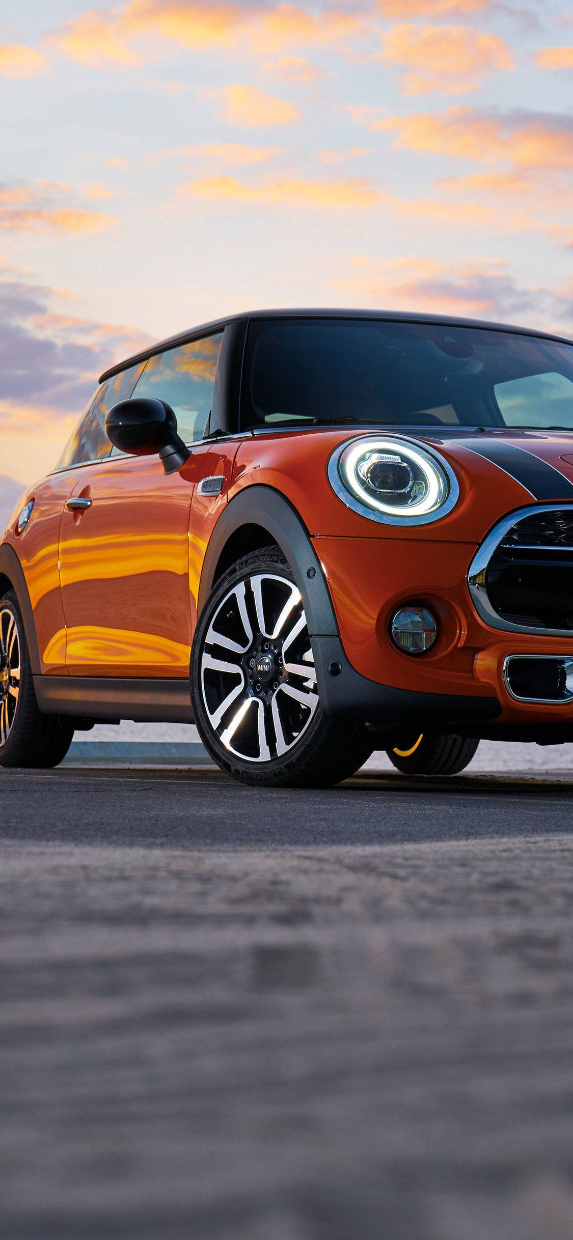 Mini Cooper 桌布 壁紙 — 一輛充滿活力的橙色MINI Cooper停泊在柏油路上，溫暖的夕陽天空下，金色雲朵點綴。